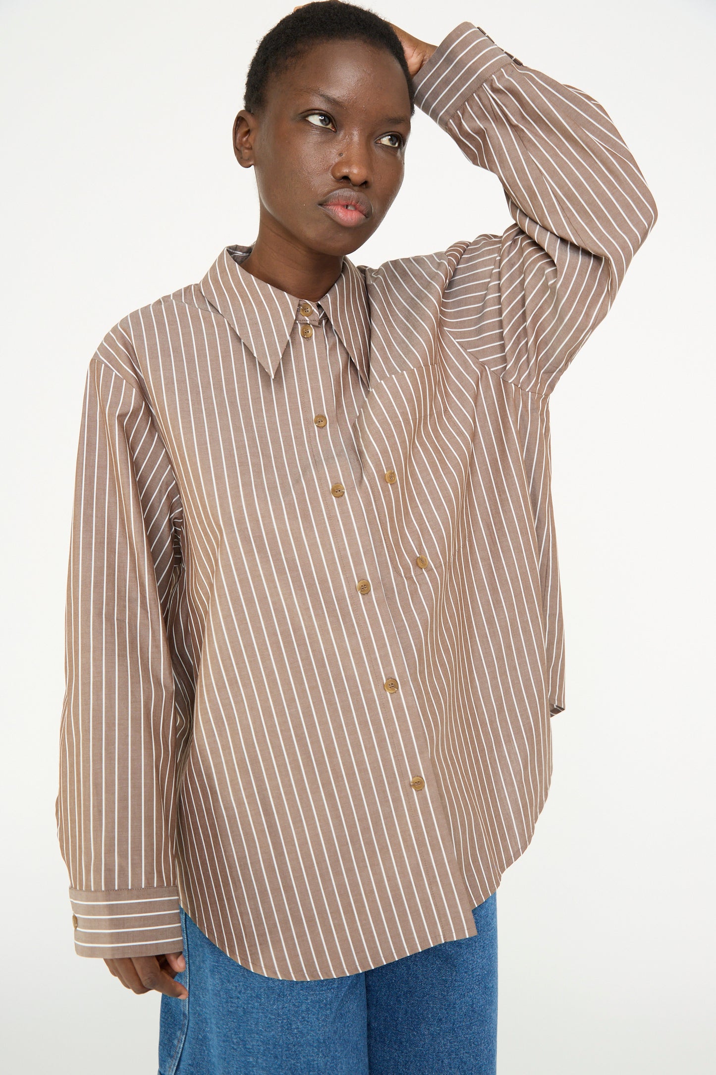 A person in an oversized Rejina Pyo Cotton Stripe Ellis Shirt in Brown and blue jeans stands against a plain background, holding their head with one hand.