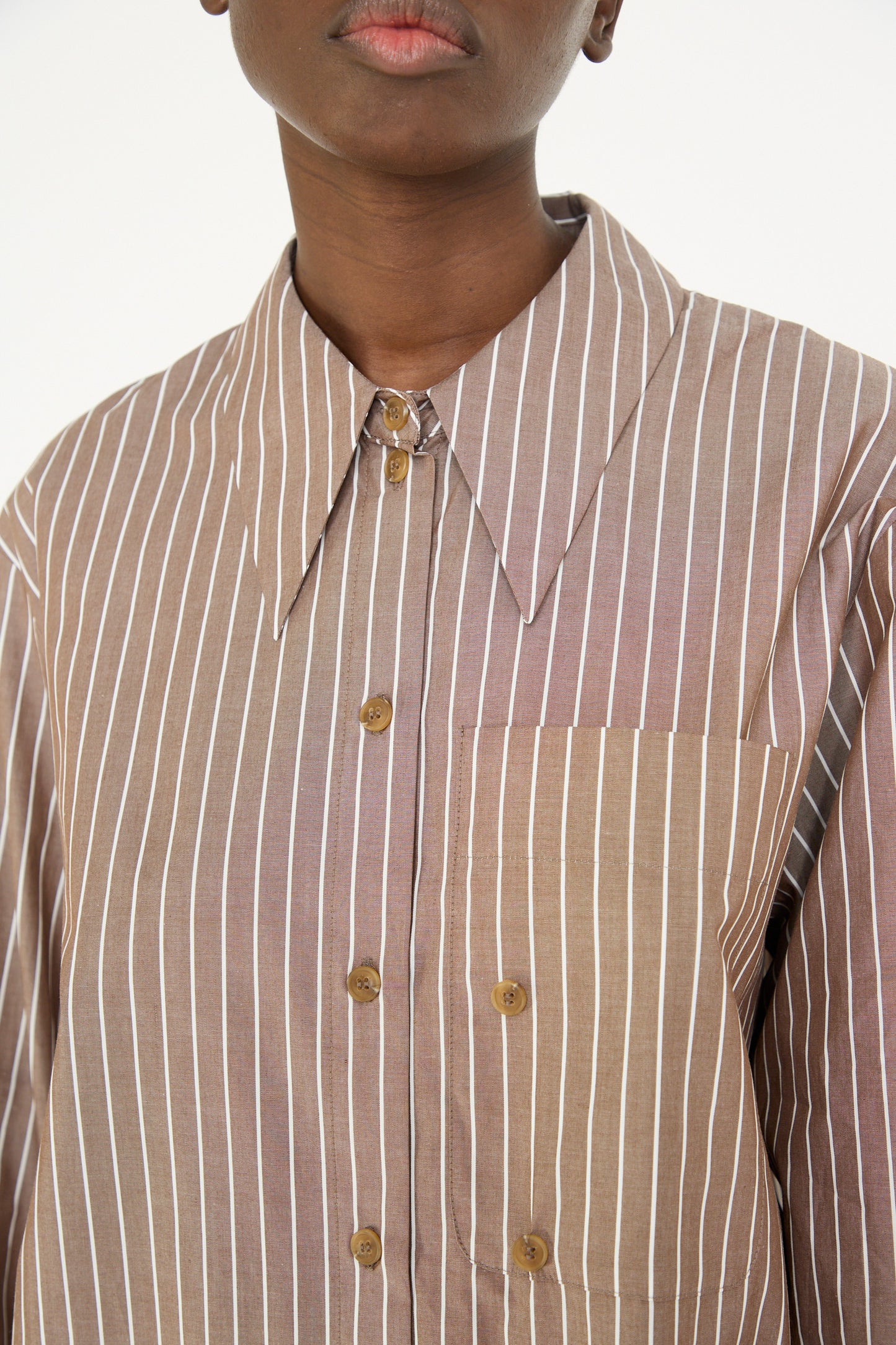 A model in the Rejina Pyo Cotton Stripe Ellis Shirt in Brown wears an oversized fit with brown and white stripes, a large pointed collar, and a convenient front pocket.