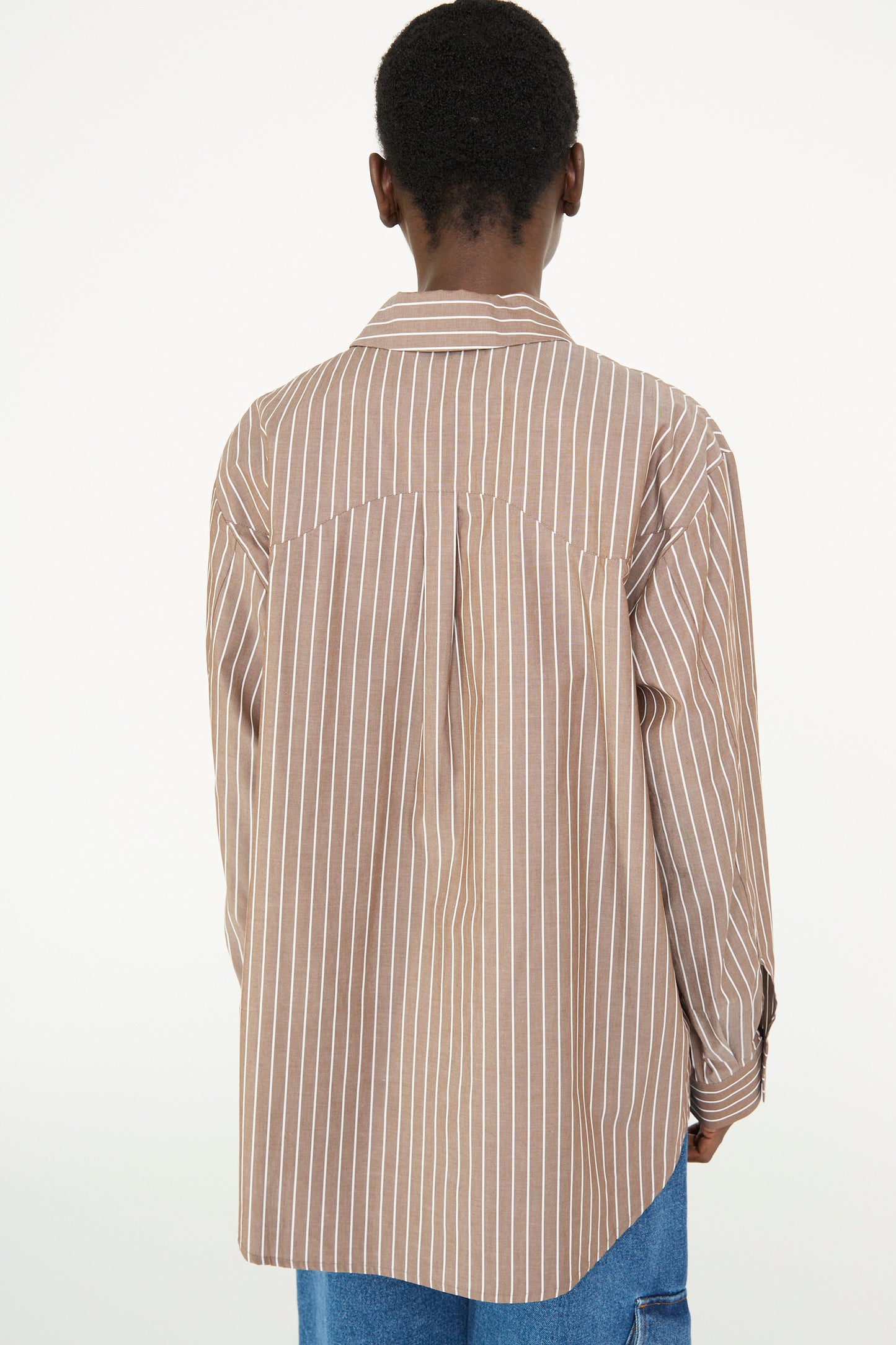 A person with short hair is seen from the back against a plain background, seamlessly blending into the minimalist setting while wearing a Rejina Pyo's oversized Cotton Stripe Ellis Shirt in Brown featuring brown and white vertical stripes.