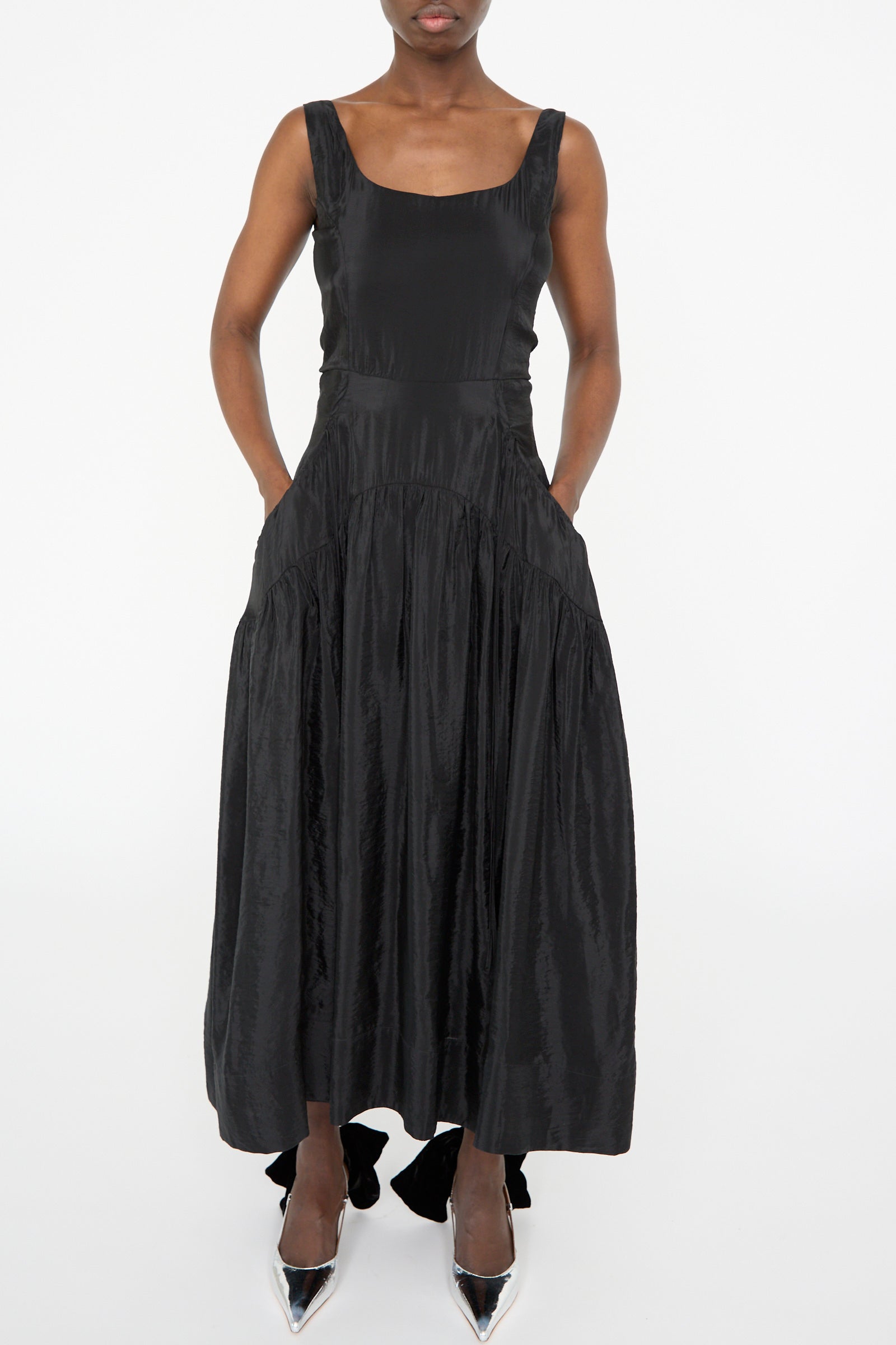 Against a plain white backdrop, a person elegantly models the Erica Dress in Black by Rejina Pyo, featuring a sleeveless design and corseted bodice, complemented by silver high-heeled shoes.