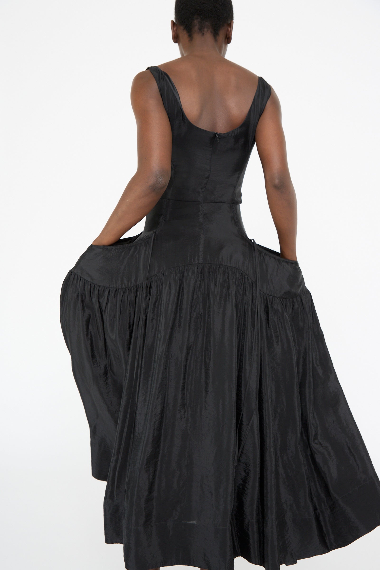 With their back to us, a person elegantly showcases the Erica Dress in Black by Rejina Pyo. This sleeveless midi dress, featuring a full skirt and subtle corseted bodice, creates an enhanced silhouette as they gently hold the sides.