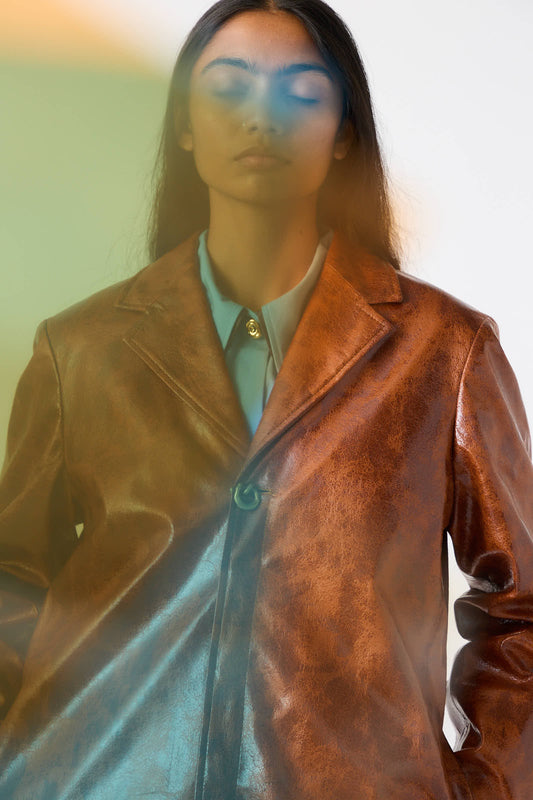 A person with long dark hair, wearing the Rejina Pyo Faux Leather Samantha Jacket in brown with a relaxed fit and a light blue shirt underneath, stands against a background of green and yellow light overlays.