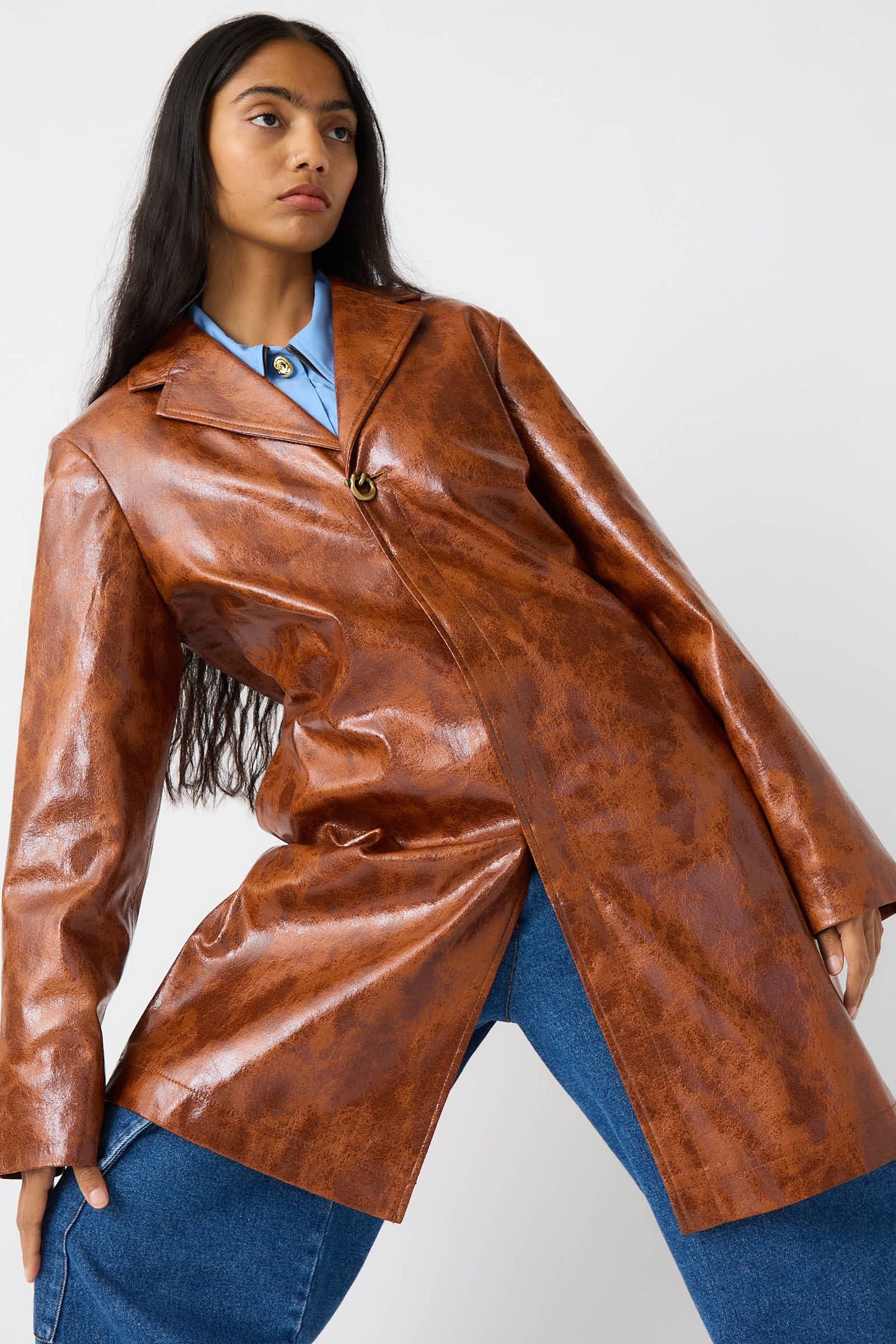 A person with long dark hair is wearing the Faux Leather Samantha Jacket in Brown by Rejina Pyo, featuring a notched collar, over a blue shirt and blue jeans, standing against a plain white background.