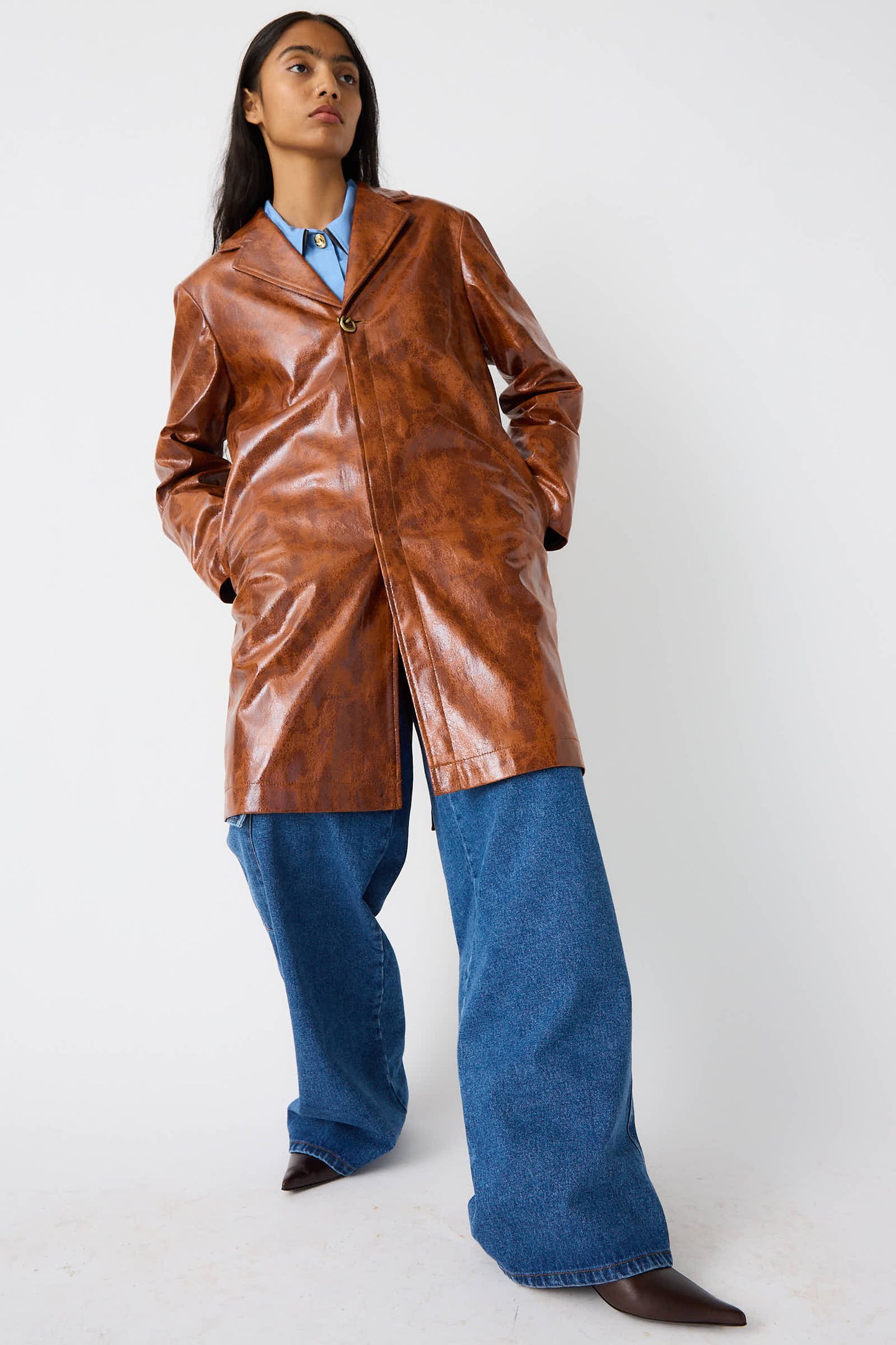 A person stands confidently in a Faux Leather Samantha Jacket in Brown by Rejina Pyo, paired with a blue shirt and wide-legged blue jeans, with hands in pockets.