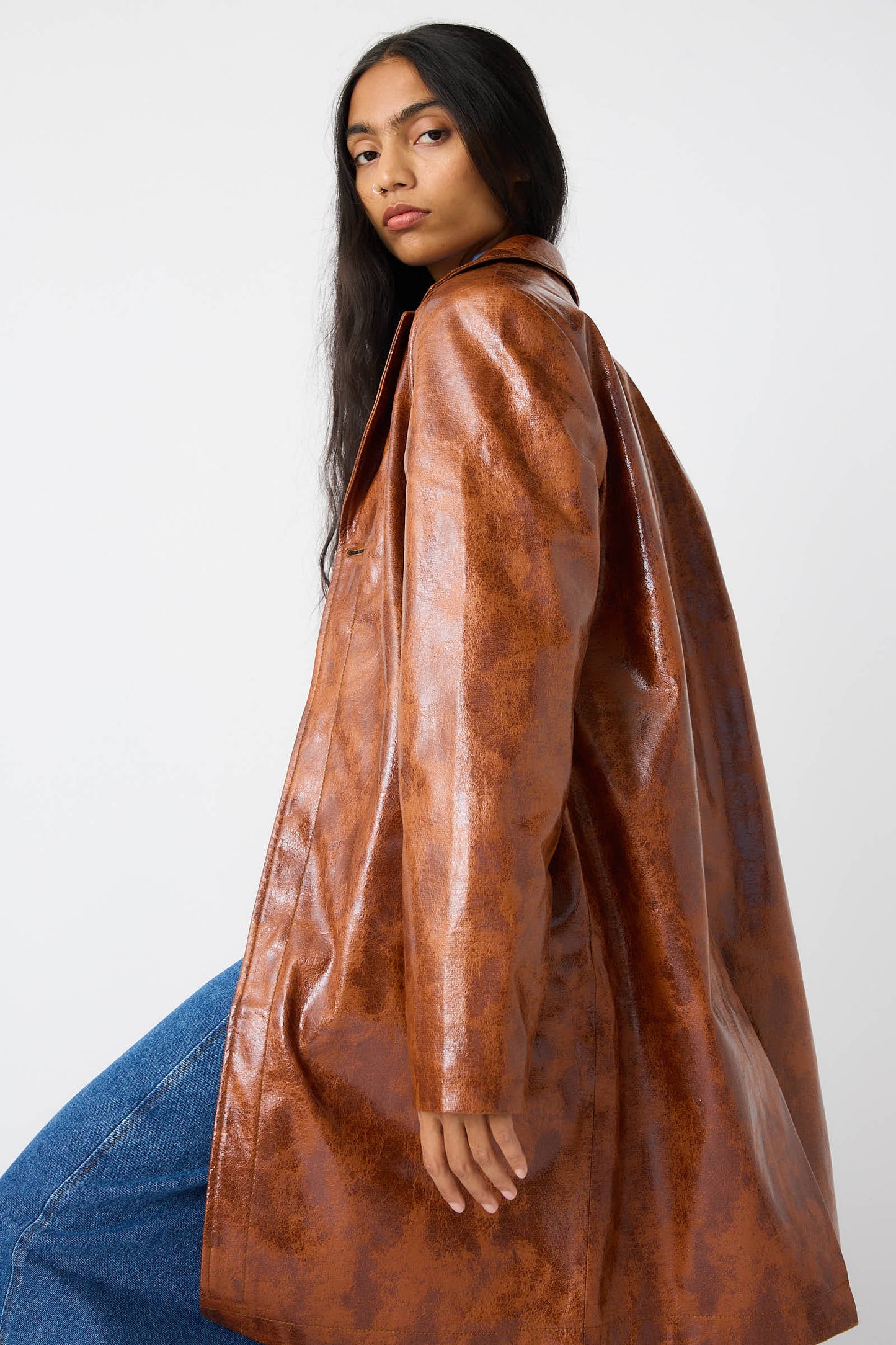 A person with long dark hair wearing the Rejina Pyo Faux Leather Samantha Jacket in Brown with a notched collar and blue jeans, looking over their shoulder against a plain background.