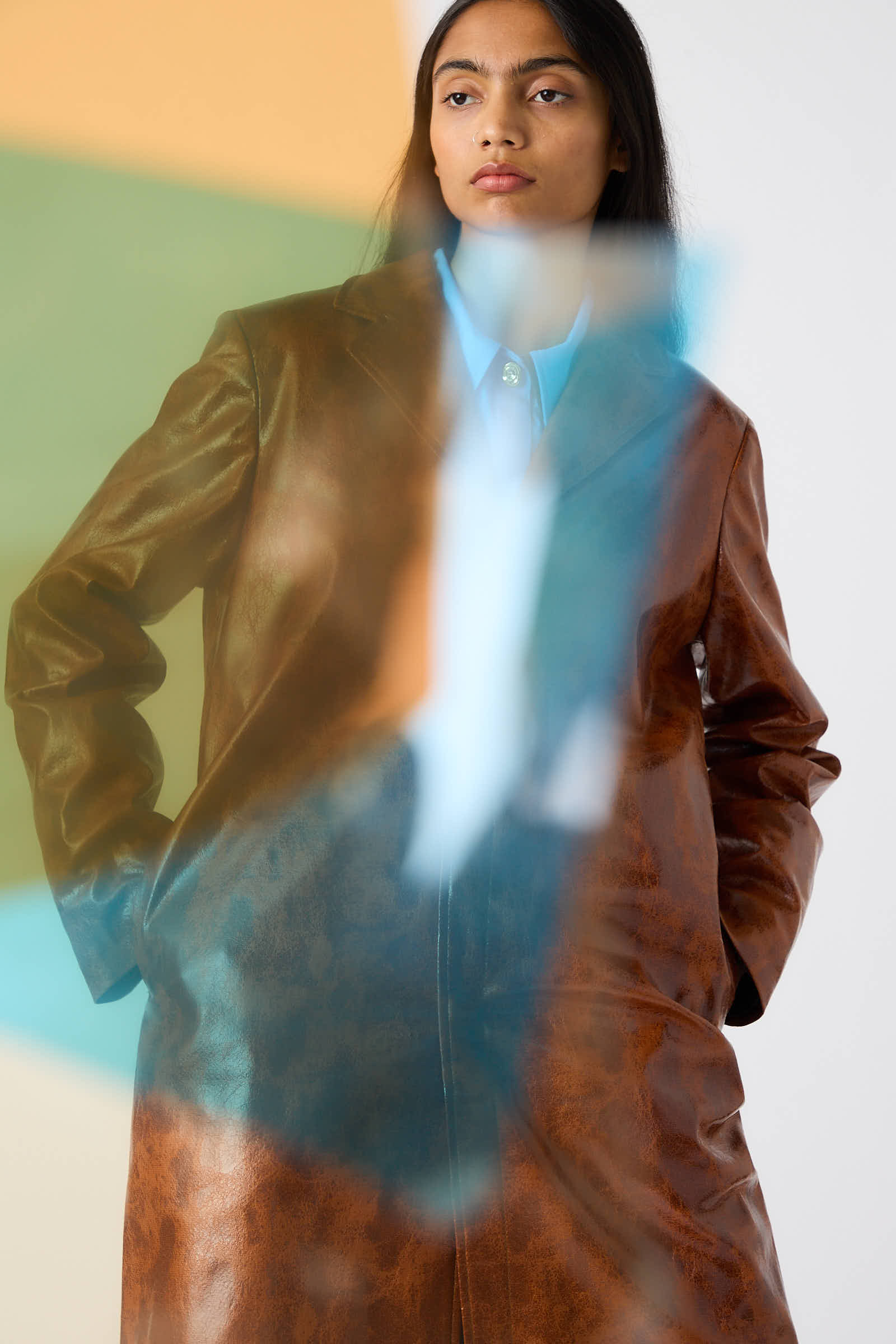 A person wearing the Rejina Pyo Faux Leather Samantha Jacket in Brown with a notched collar looks to the side. The image features colorful blurred elements in the foreground, adding depth and texture to the scene.