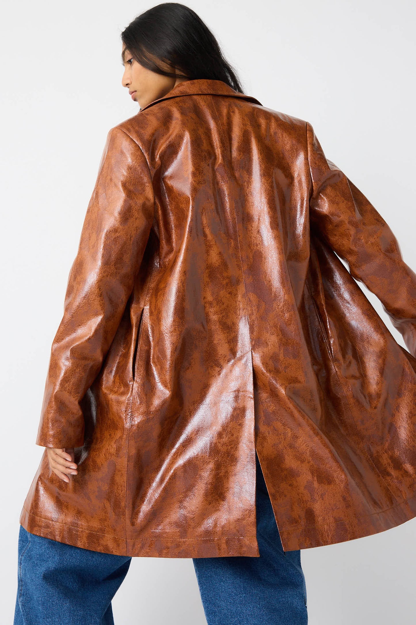 A person with long dark hair is turned away, wearing the Rejina Pyo Faux Leather Samantha Jacket in Brown, which features a notched collar and a slit at the back, paired with relaxed fit blue jeans.