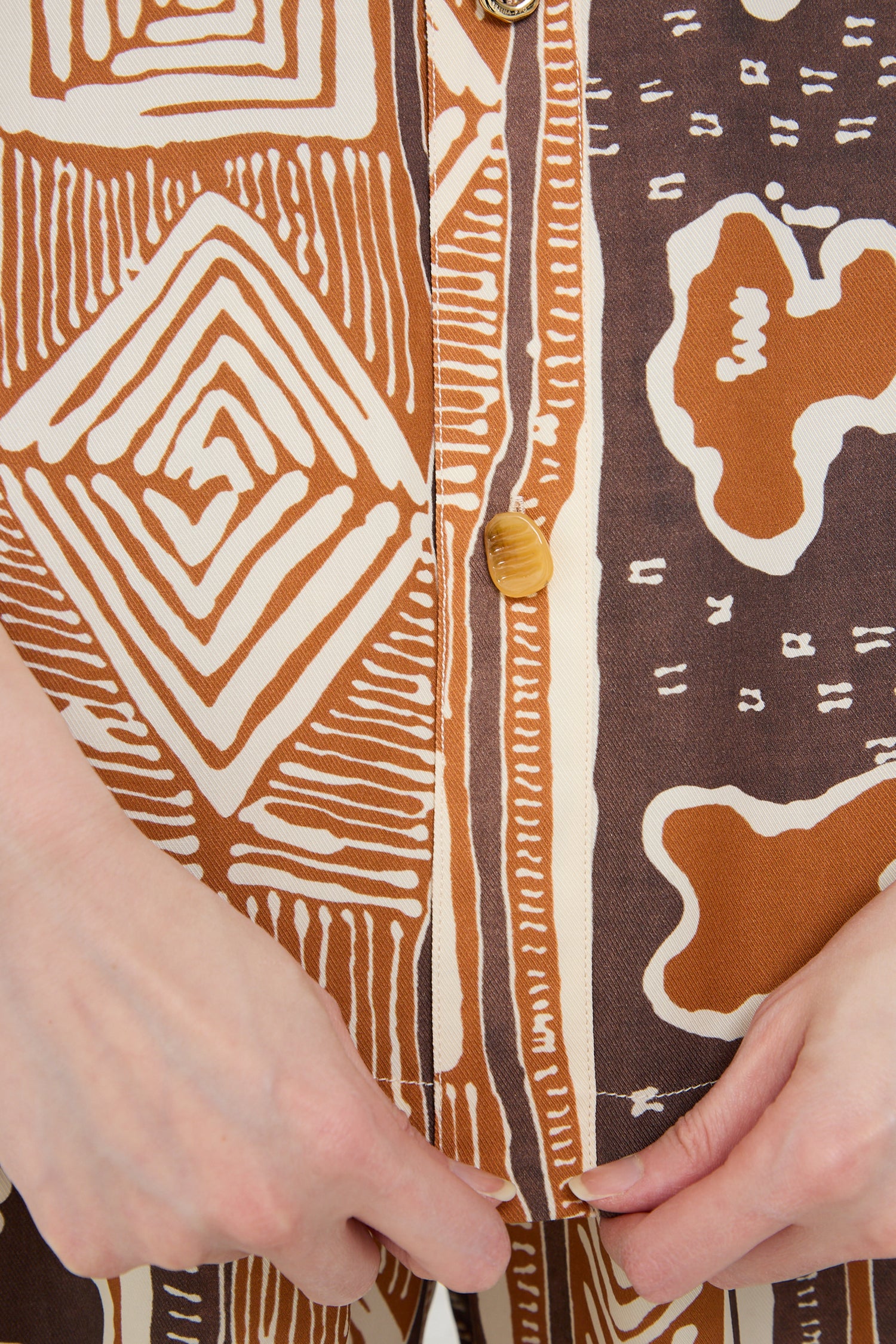 Close-up of a person's hands zipping up a Rejina Pyo Marty Shirt in Print Brown with a hand-painted print design and a visible yellow button.