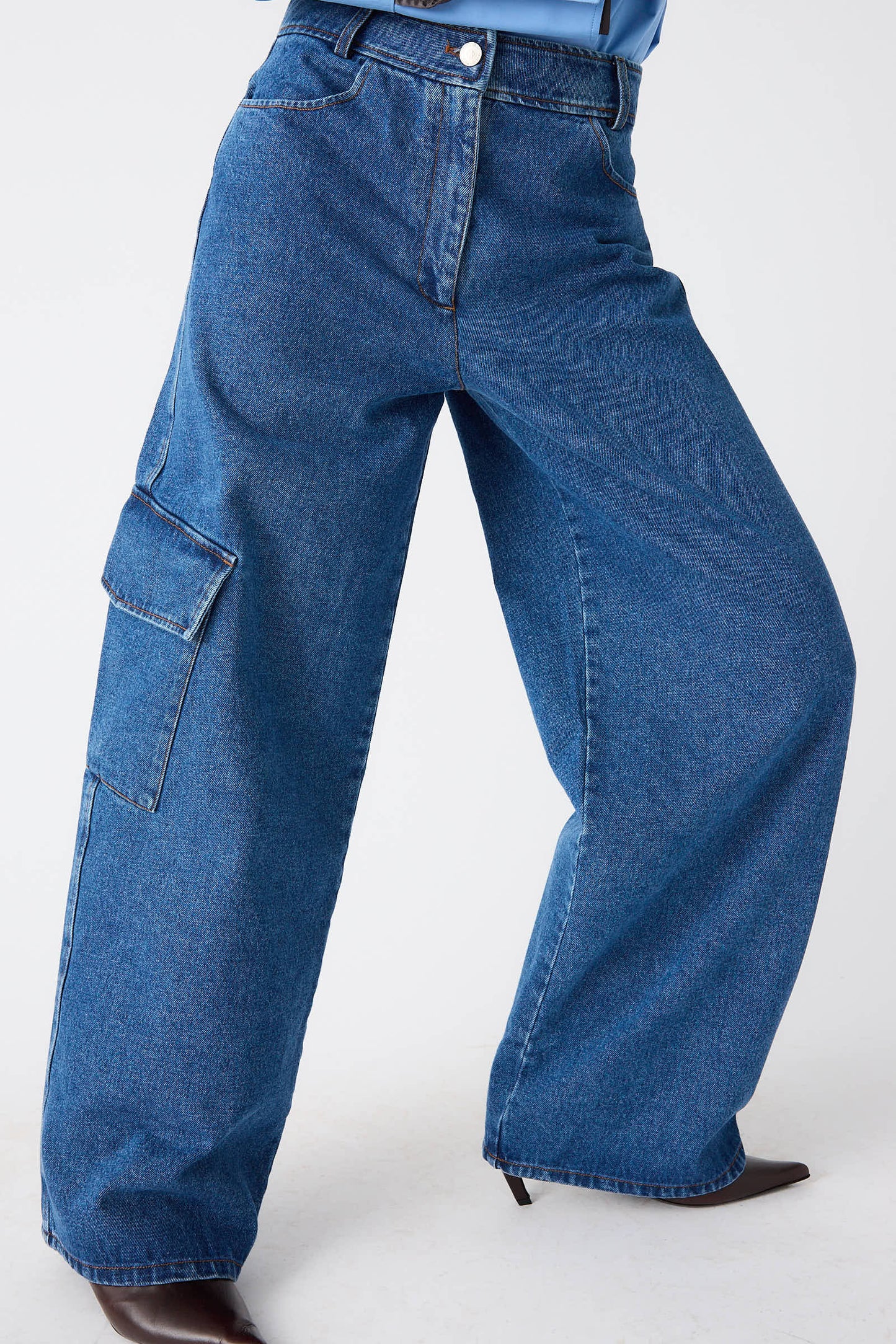 A person wearing Rejina Pyo's Organic Cotton Emery Jeans in Denim Blue, featuring a mid-rise and wide-leg design with a side cargo pocket, paired with a light blue shirt and black pointed shoes.