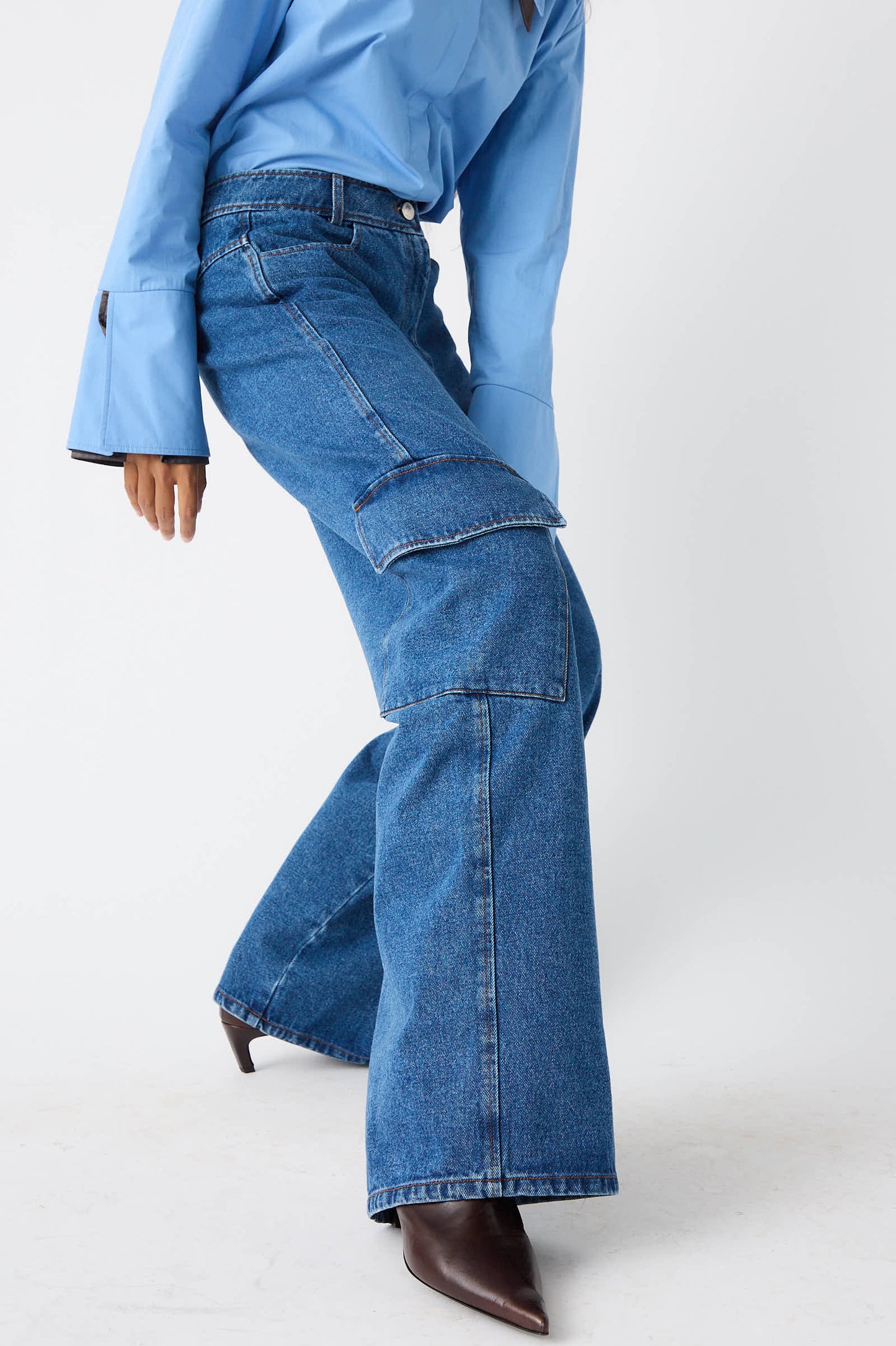 A person wearing a light blue shirt and Rejina Pyo's Organic Cotton Emery Jeans in Denim Blue, featuring mid-rise wide legs and large pockets, bends slightly while standing on a white background.