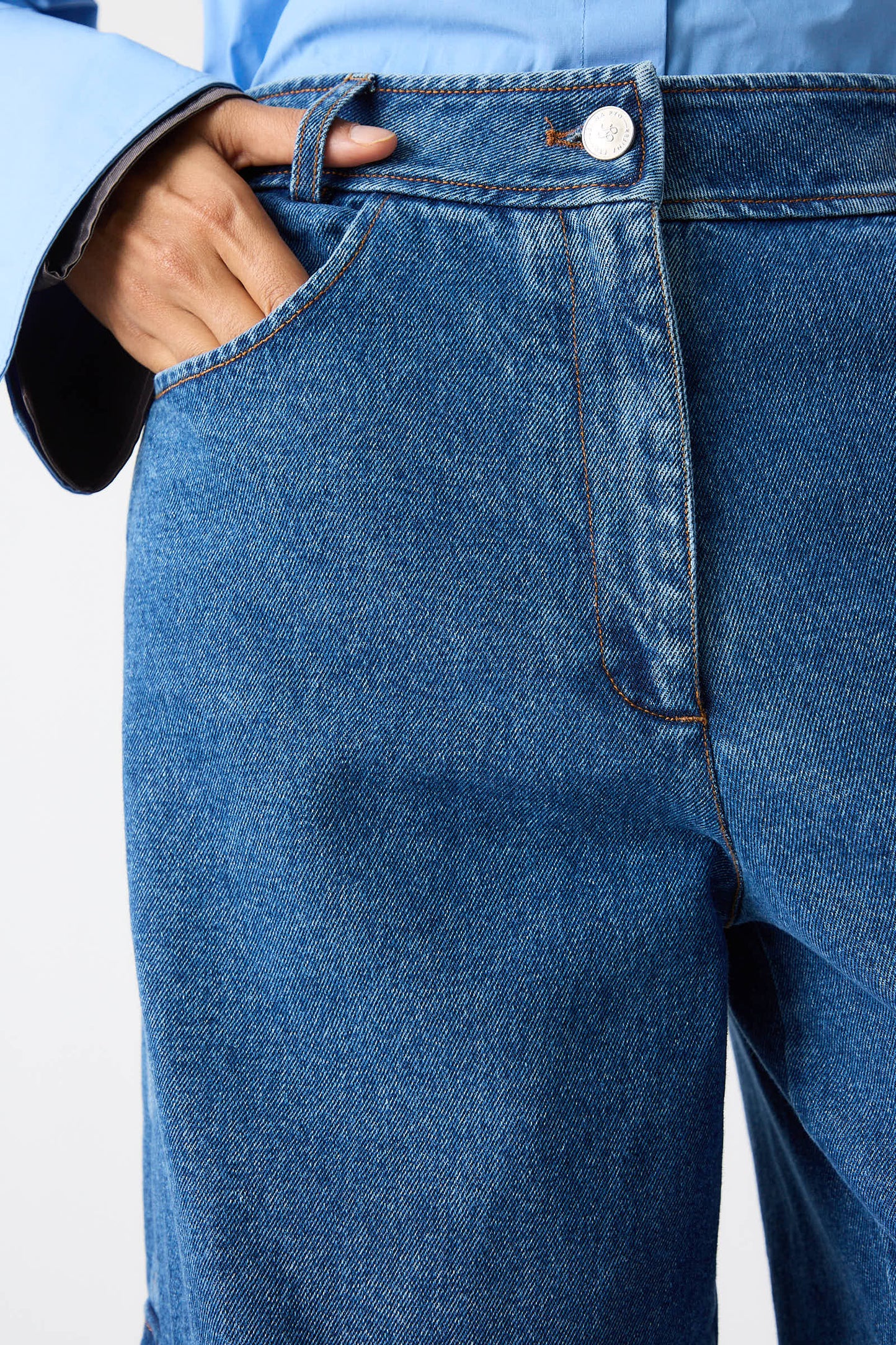 Close-up of a person wearing Rejina Pyo's Organic Cotton Emery Jeans in Denim Blue paired with a light blue shirt. One hand is partially tucked into a front pocket of the jeans, which feature an organic cotton blend for a comfortable fit.