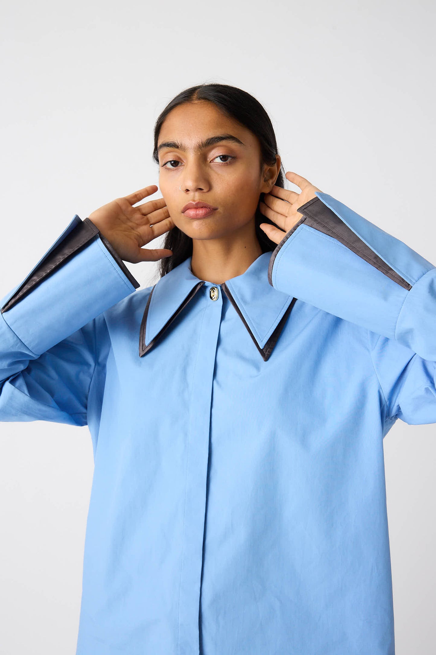 A person in the Rejina Pyo Organic Cotton Parker Shirt in Blue, a relaxed fit, long-sleeve button-down design with black trim on the collar and cuffs, holding their hands near their neck against a plain background.