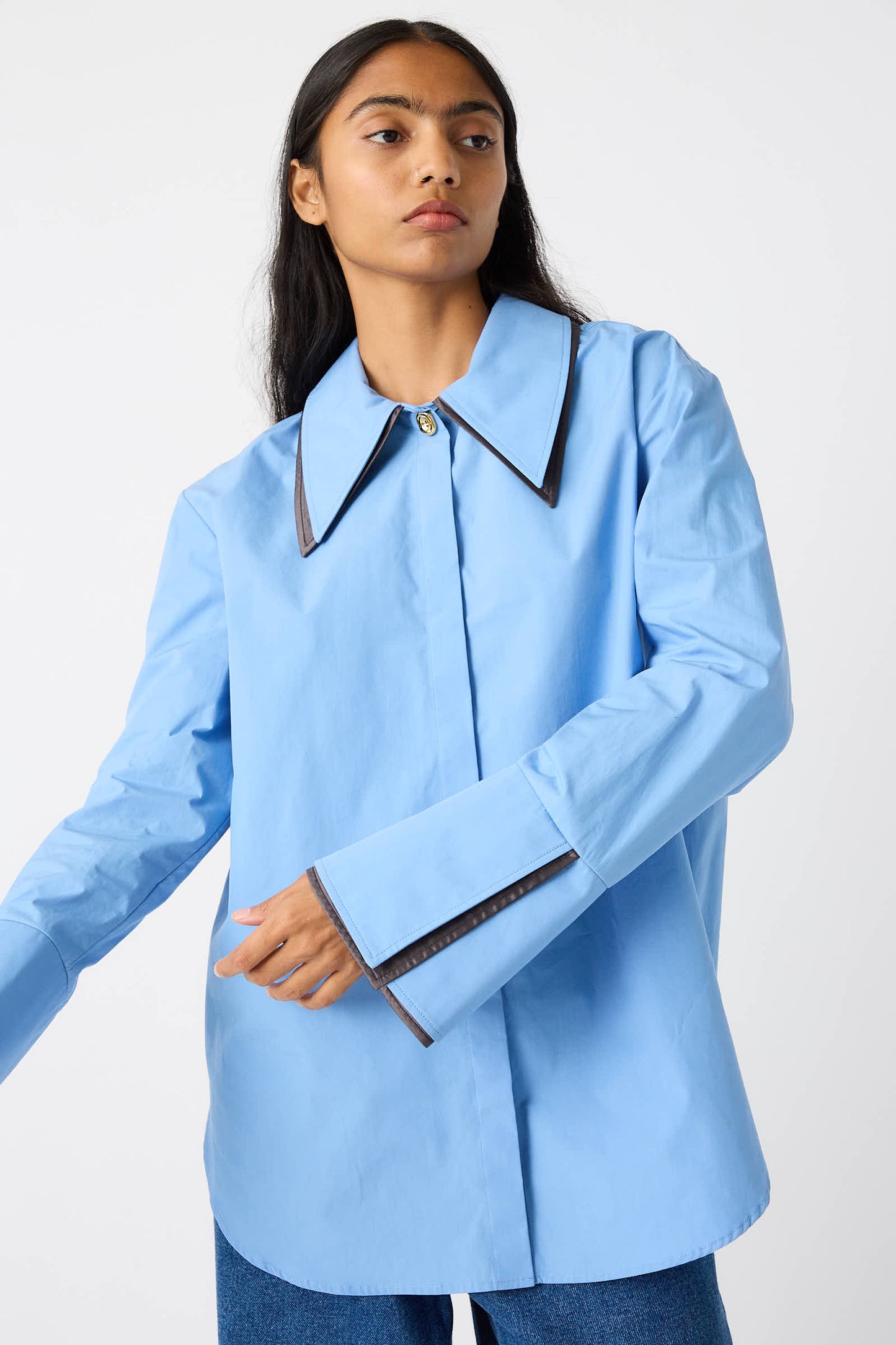 A person with long hair wearing the Rejina Pyo Organic Cotton Parker Shirt in Blue, which features a large collar and dark trim, is standing against a plain background.