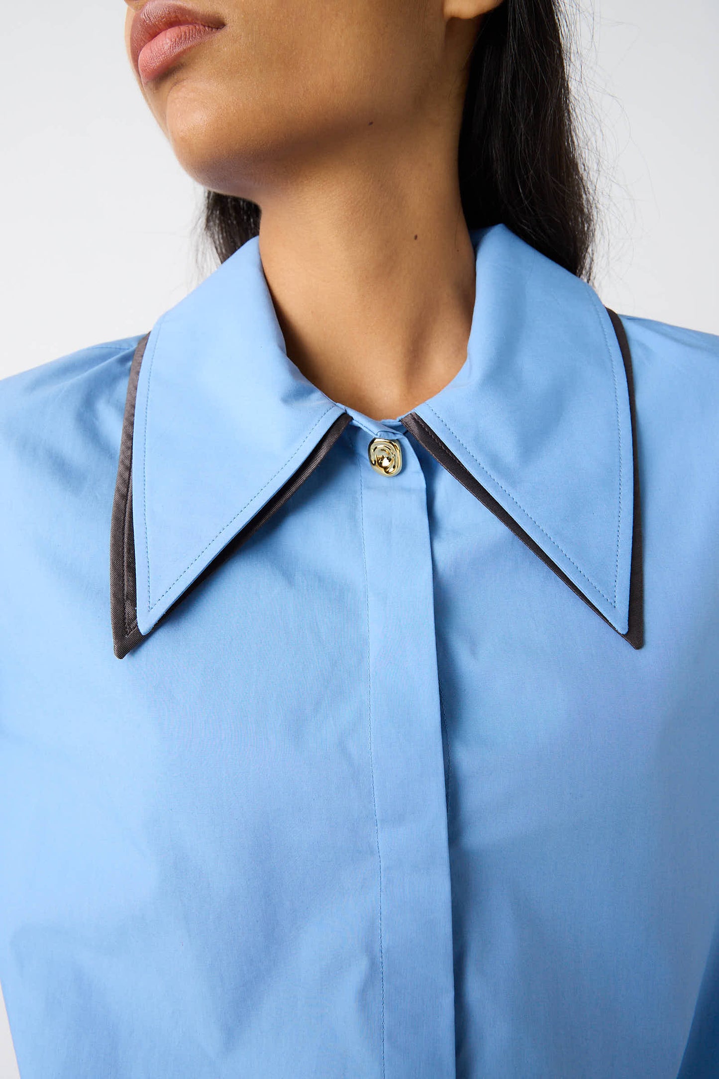 A person wearing a relaxed fit Rejina Pyo Organic Cotton Parker Shirt in Blue, buttoned at the neck, featuring black trim on the edges of the collar.