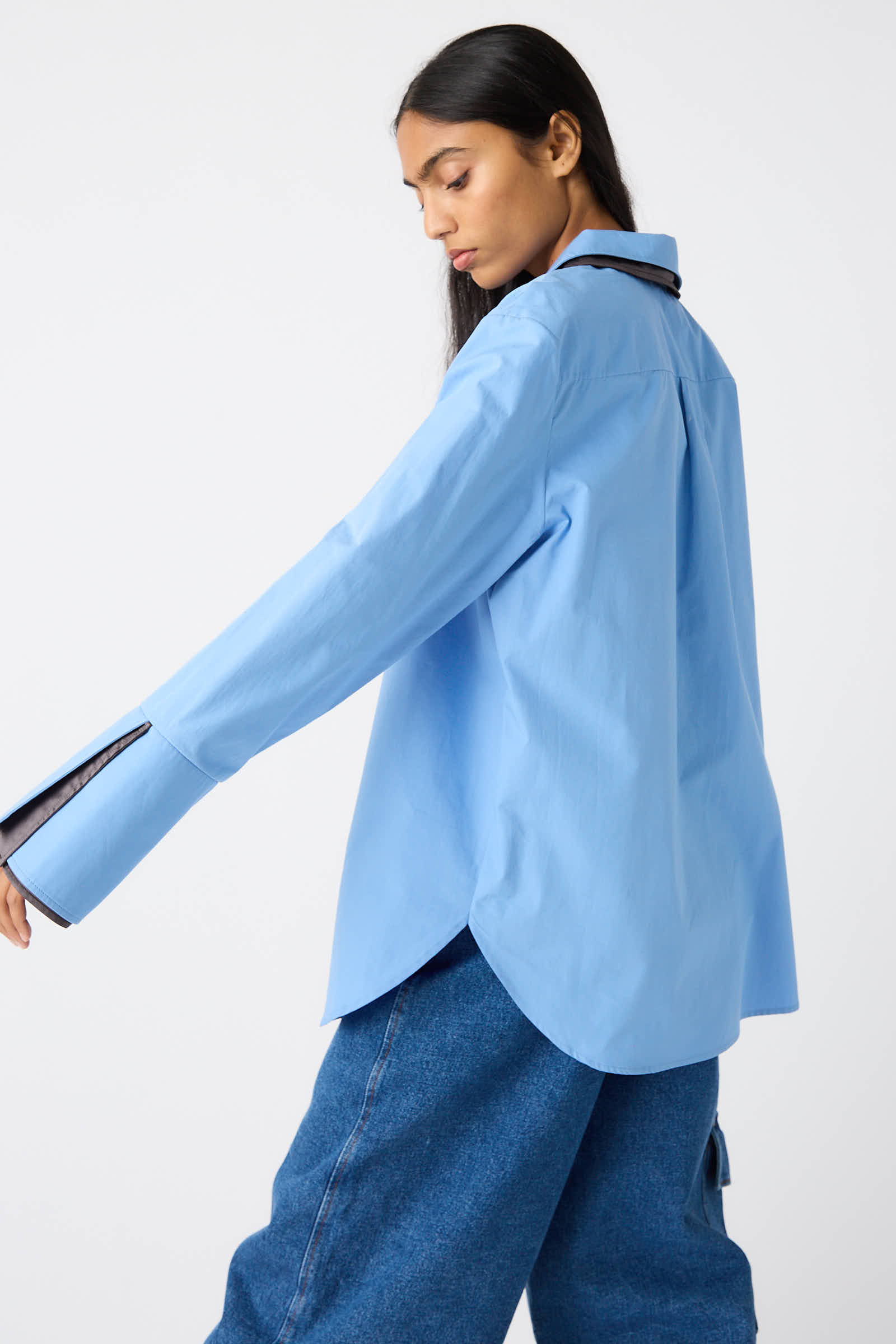 Person wearing a Rejina Pyo Organic Cotton Parker Shirt in Blue with relaxed fit and black trim, paired with loose-fitting jeans, stands against a white background, looking down and to the side.