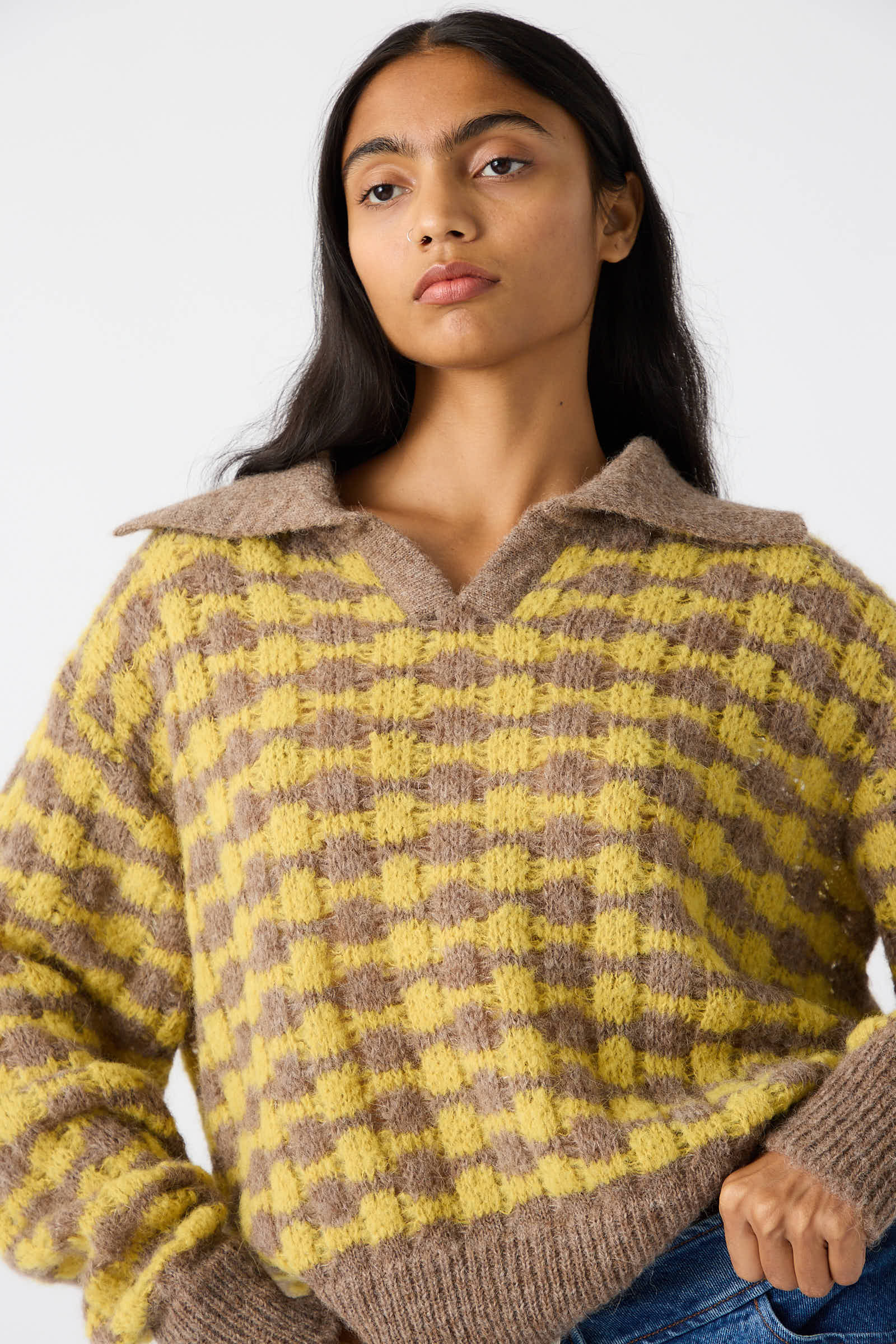 A person with long dark hair wearing a Rejina Pyo Rae Sweater in Yellow, featuring a relaxed fit and a yellow and brown checkered alpaca blend, stands against a plain background.