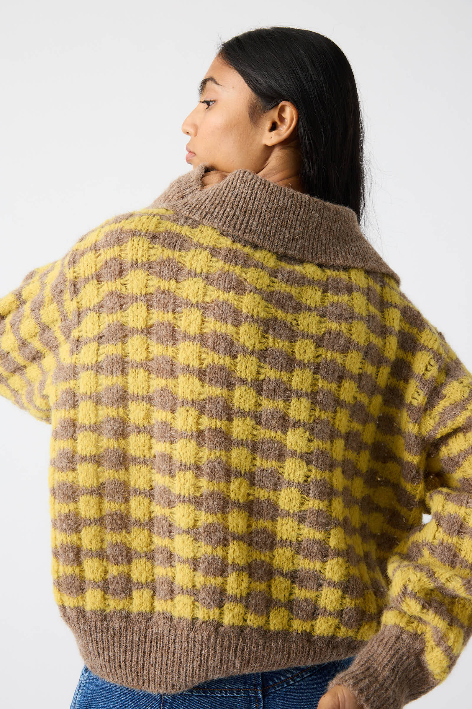 A person with long hair is seen from the back, wearing the Rae Sweater in Yellow by Rejina Pyo, characterized by its yellow and brown plaid knit design and thick collar. The relaxed fit of its alpaca blend fabric enhances its cozy appeal.