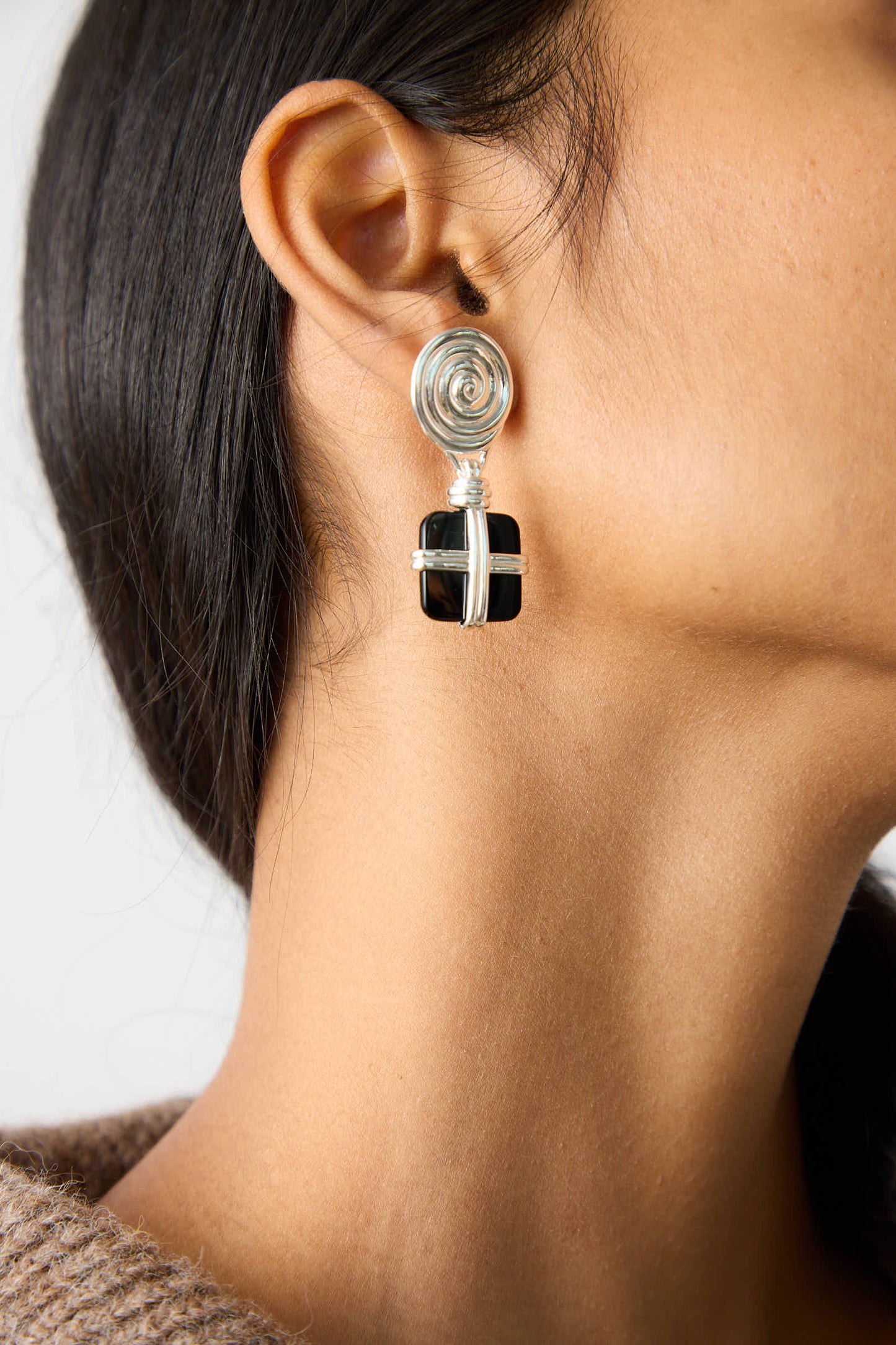 A close-up image of an ear adorned with the lightweight Silver Plated Squiggle Earrings with Black Onyx by Rejina Pyo, featuring a silver and black geometric design shaped like a wrapped gift box, complemented by a silver plated brass spiral stud.