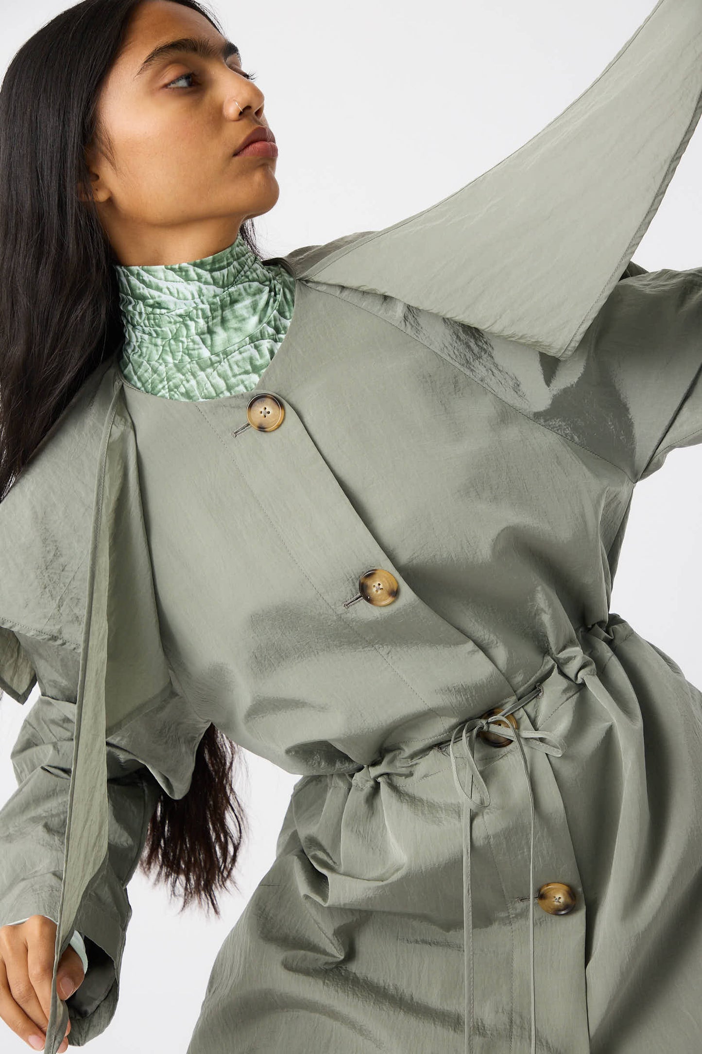 A person wearing the Rejina Pyo Taffeta Alana Trenchcoat in Grey, featuring oversized dimensions and large buttons, poses against a plain background. They also sport a high-neck, textured green top and have long, dark hair.