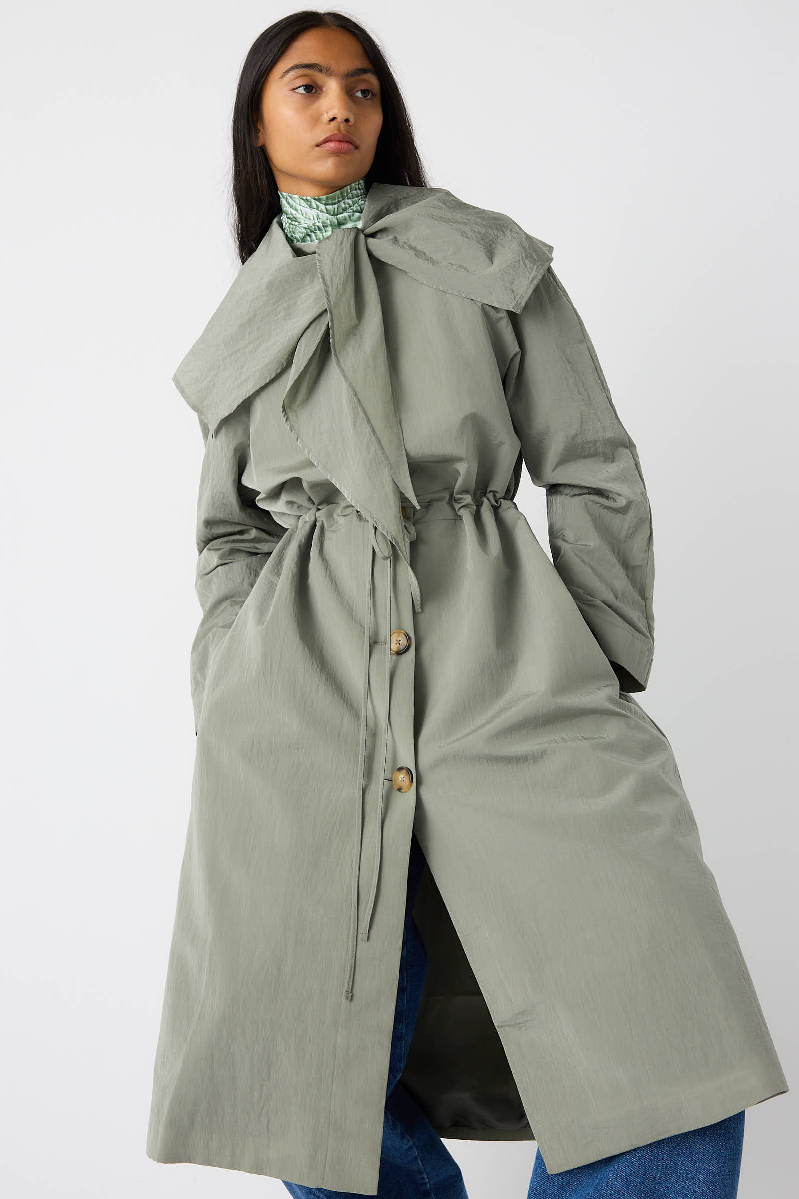 Person wearing the Rejina Pyo Taffeta Alana Trenchcoat in Grey, featuring an oversized self-tie collar and a cinched waist, standing against a plain white background.