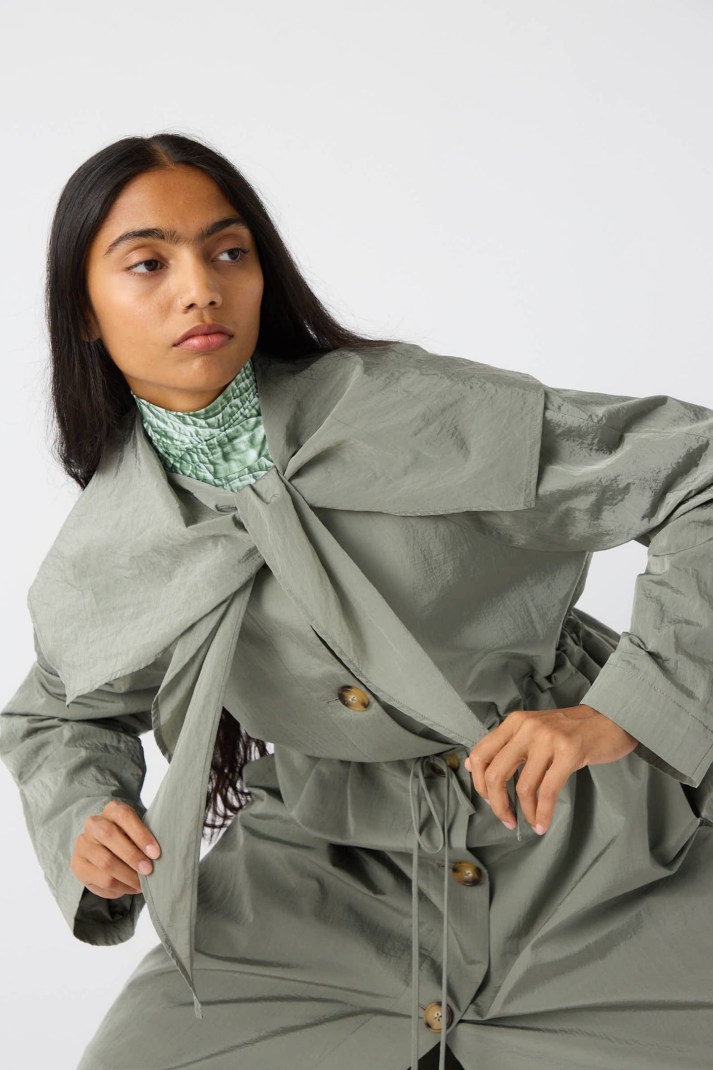 A woman with long black hair wears the Rejina Pyo Taffeta Alana Trenchcoat in grey, featuring an oversized self-tie collar and a cinched waist, posing with one hand on the coat's tie and the other resting on her hip, set against a plain white background.