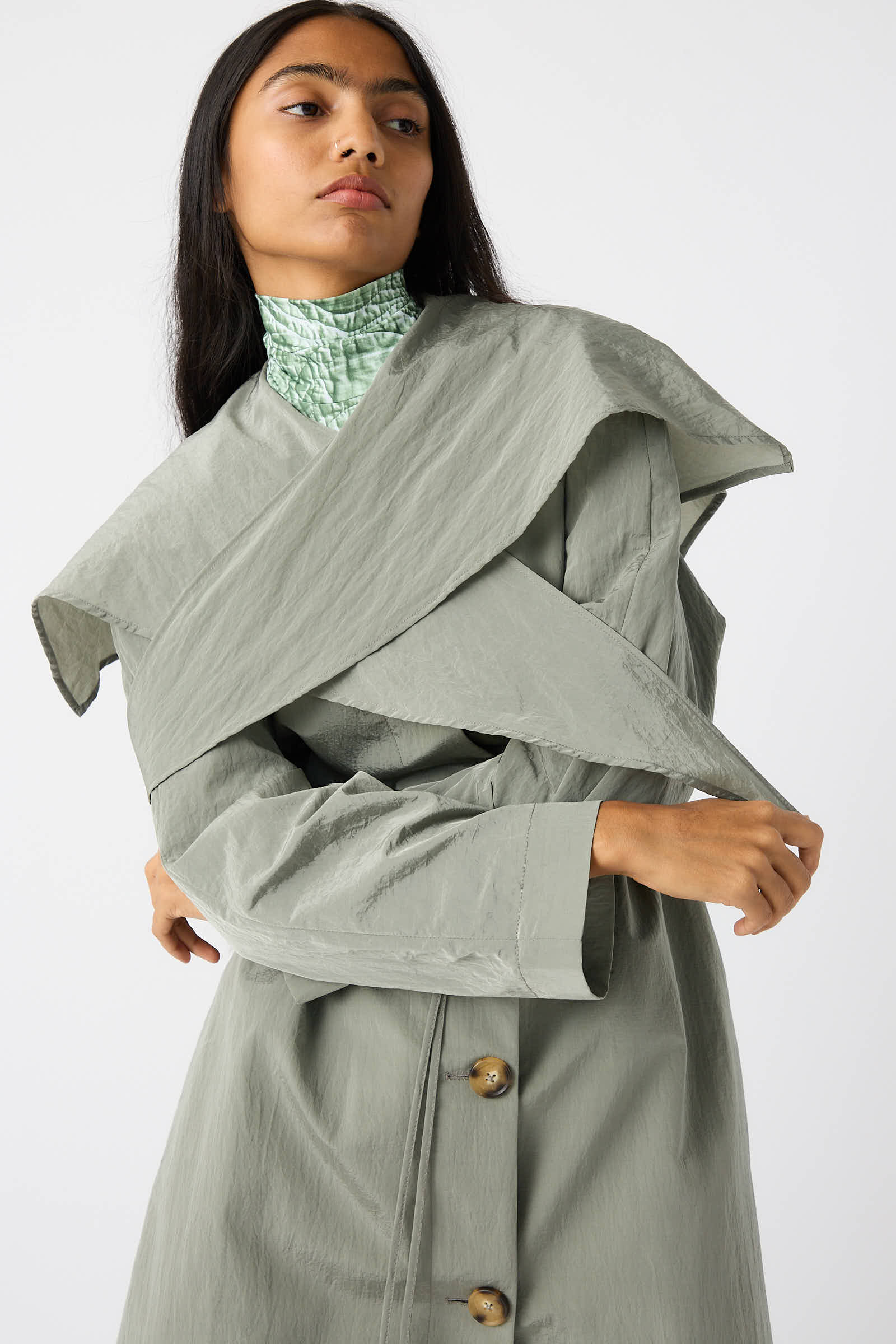 A person wearing a Taffeta Alana Trenchcoat in Grey by Rejina Pyo, with three visible buttons and a textured finish, poses with arms partially crossed. Underneath the trench coat, they sport a high-necked, patterned top featuring an oversized self-tie collar.