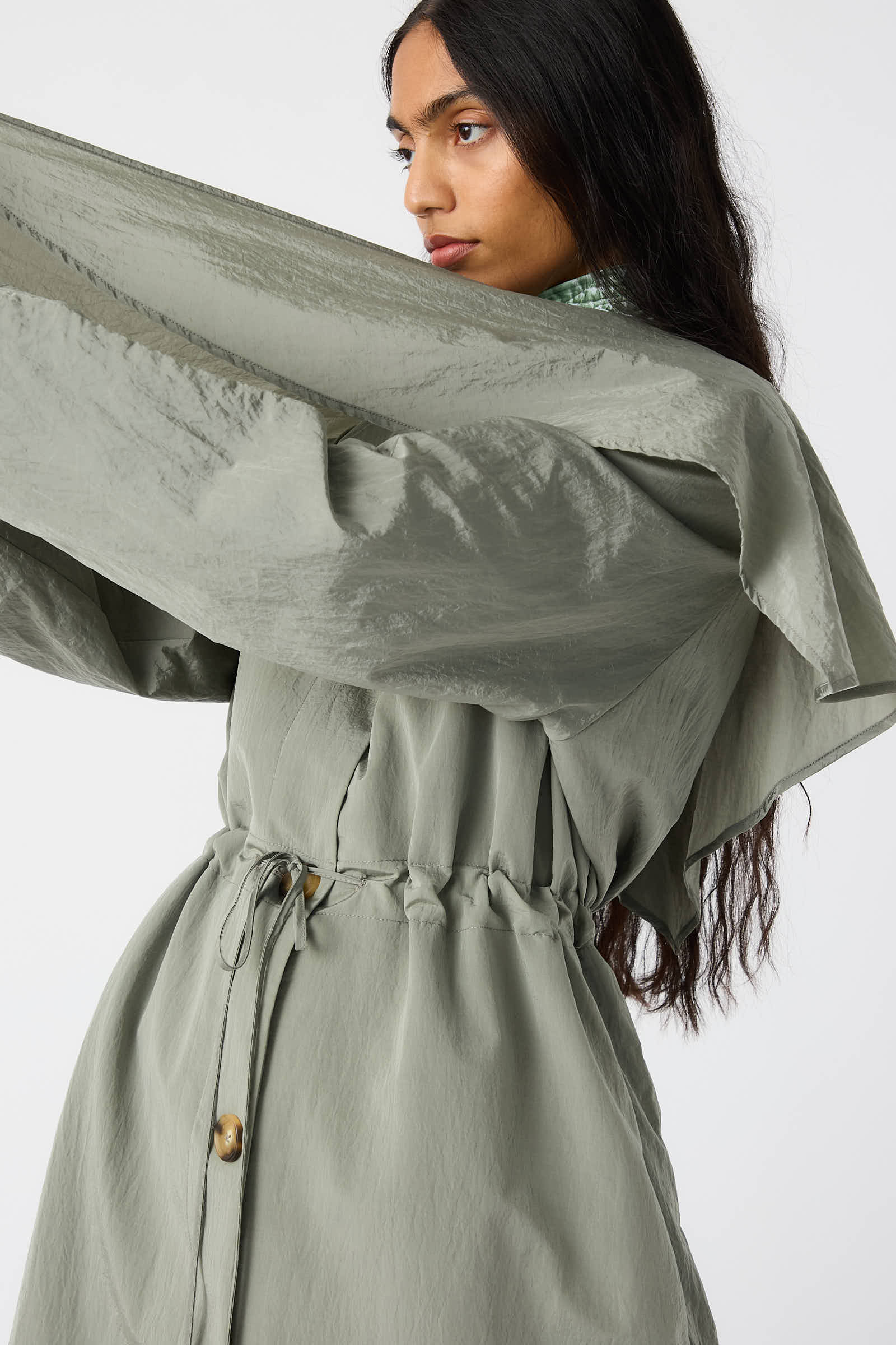 A person with long dark hair wears the grey Taffeta Alana Trenchcoat by Rejina Pyo, which features an elegant cinched waist, a high collar, and button details against a plain white background.