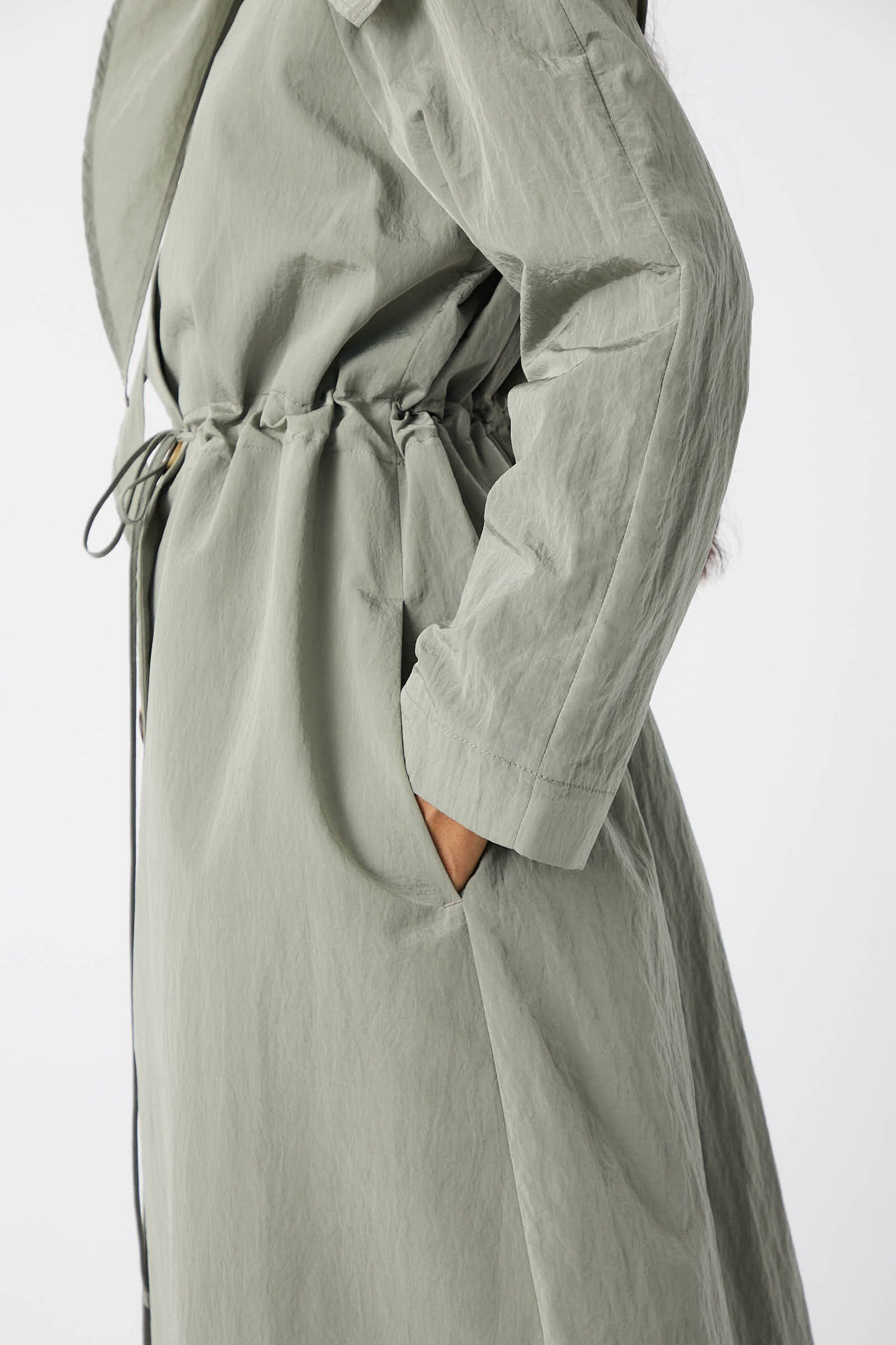 Side view of a person wearing the Taffeta Alana Trenchcoat in grey by Rejina Pyo, featuring an oversized self-tie collar and a cinched waist, with their right hand in its pocket.