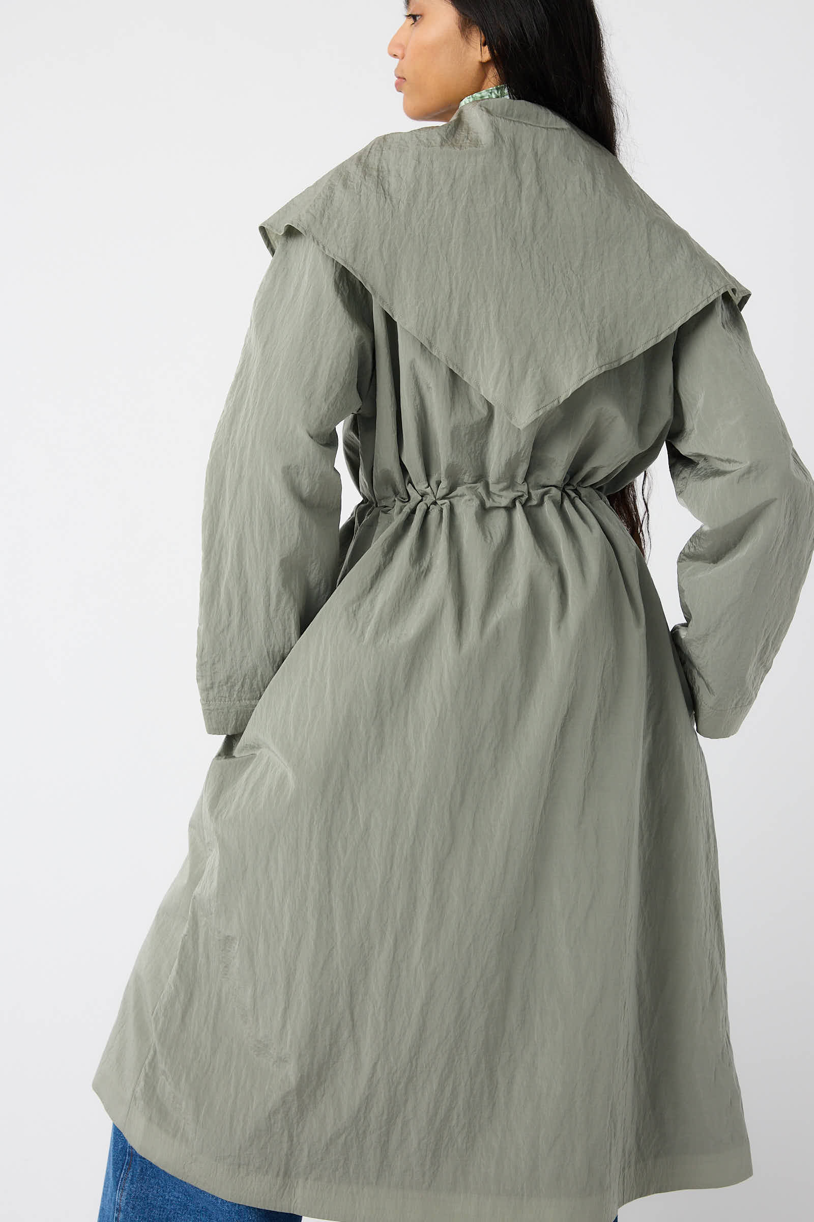 A person with long hair is seen from the back, wearing the Rejina Pyo Taffeta Alana Trenchcoat in Grey, featuring a long-sleeved design with a cinched waist and an oversized self-tie collar.