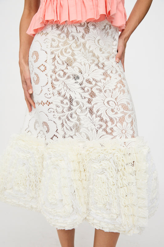 Someone wearing the Renata Brenha Acarajé Skirt in Tonal White—a white lace midi with floral patterns and ruffled hem—paired with a pink pleated top.