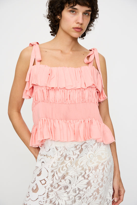 A woman wears the Renata Brenha Babado Top in Coral Pink, featuring ruffles and an open back with shoulder ties, styled with a white lace skirt against a plain background.