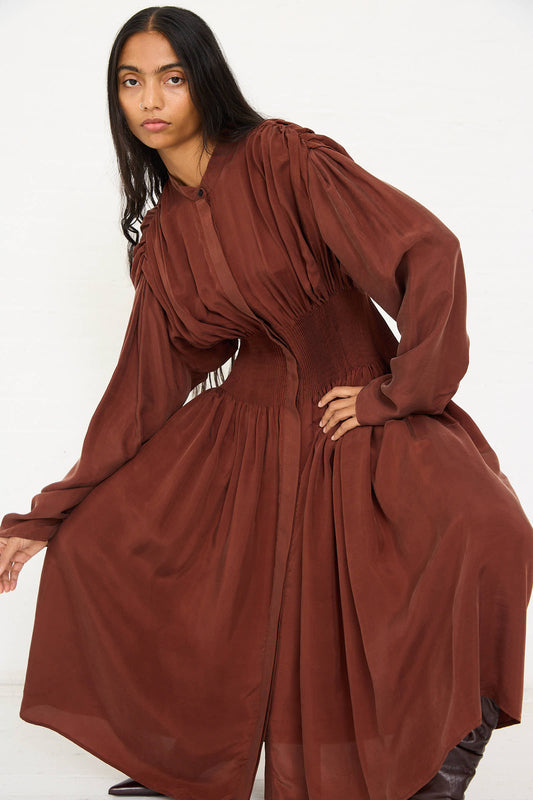 A person with long, dark hair wearing the Renata Brenha Entre Rios Dress in Washed Brown, a loose maxi dress made from recycled materials and featuring gathered fabric with a fitted waist corset, stands against a plain white background, looking at the camera.