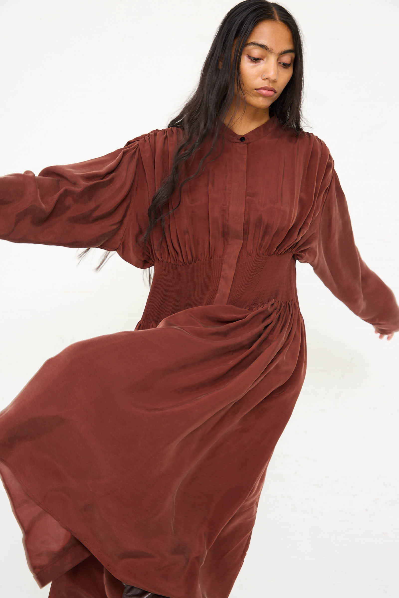 A woman with long dark hair twirls in the flowing, silky feel of the Entre Rios Dress in Washed Brown by Renata Brenha, made from recycled materials, against a plain background.