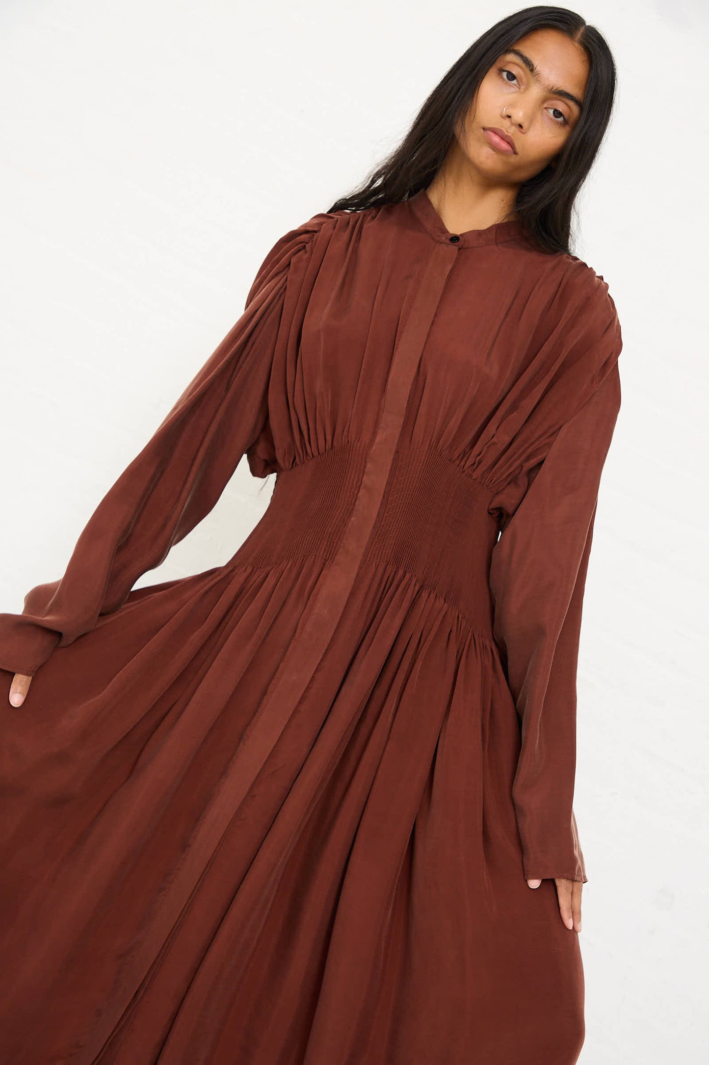 A person with long dark hair wearing the Renata Brenha Entre Rios Dress in Washed Brown, a flowing, long-sleeved maxi dress made of recycled materials, stands against a plain background.