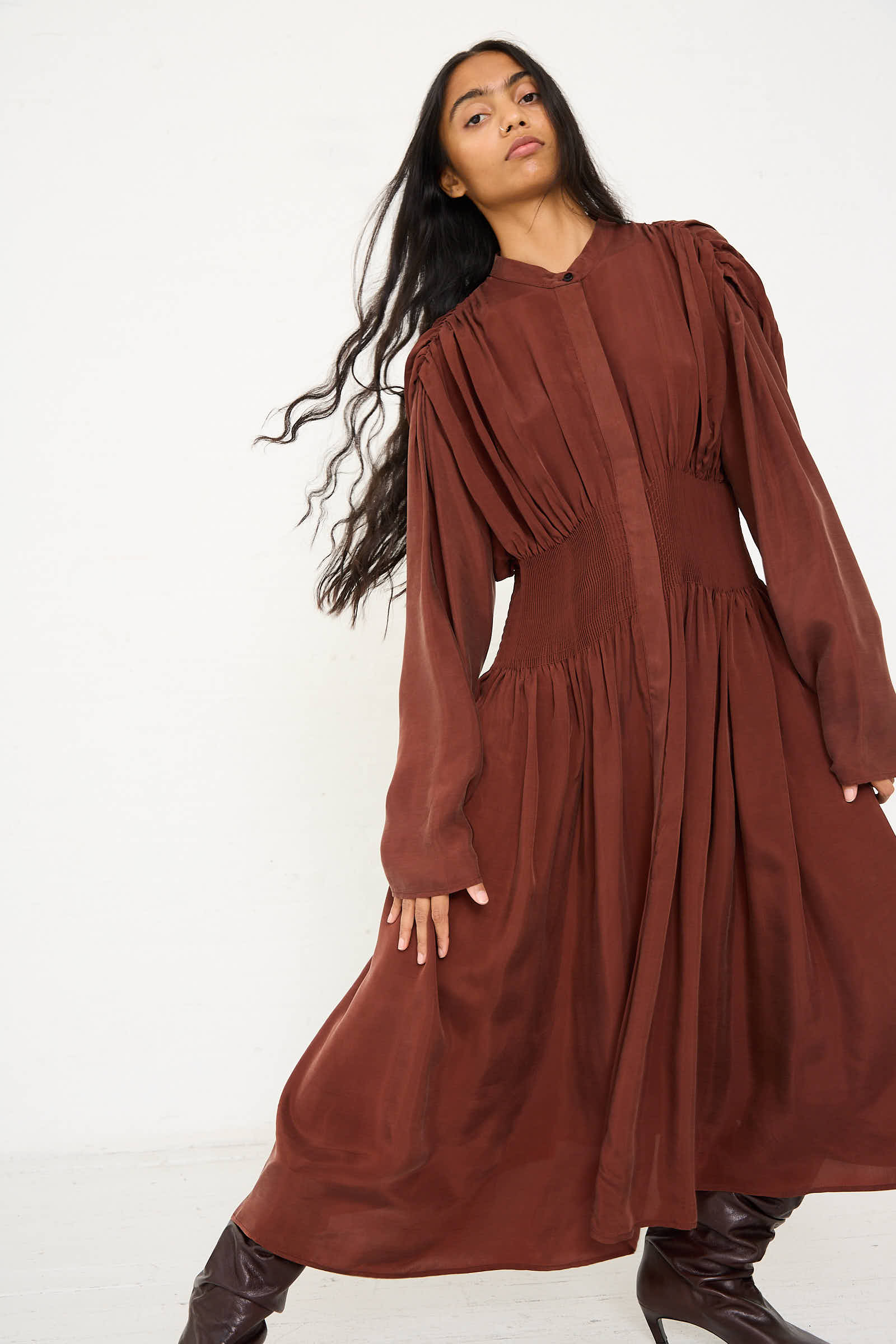 A person with long hair, wearing the Renata Brenha Entre Rios Dress in Washed Brown—a flowing, long-sleeved maxi dress made from recycled materials—stands against a plain background with one arm bent slightly.