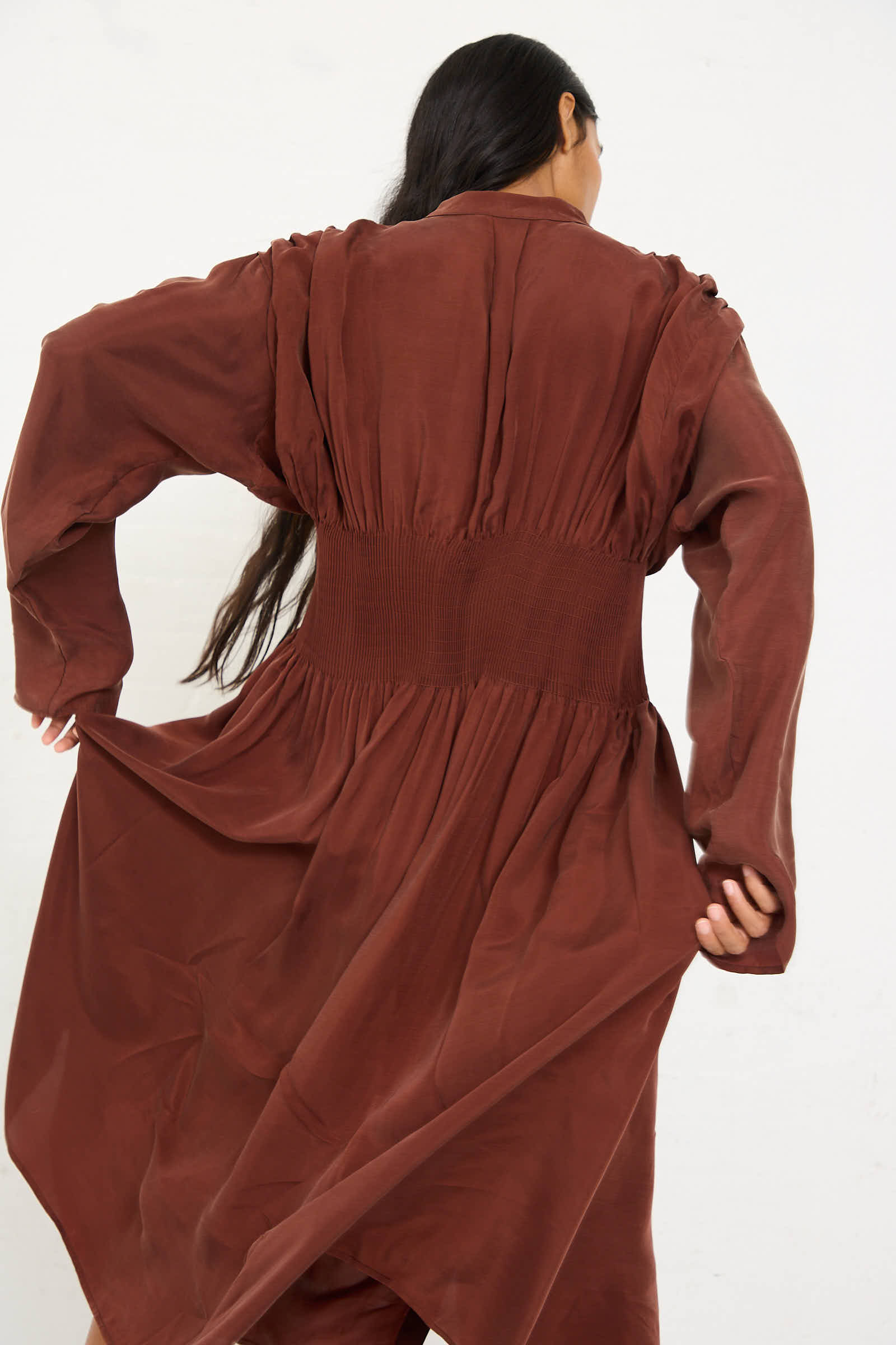 A person with long, dark hair is seen from the back wearing the loose-fitting Entre Rios Dress in Washed Brown by Renata Brenha, which features gathered detailing at the waist for a silky effect.