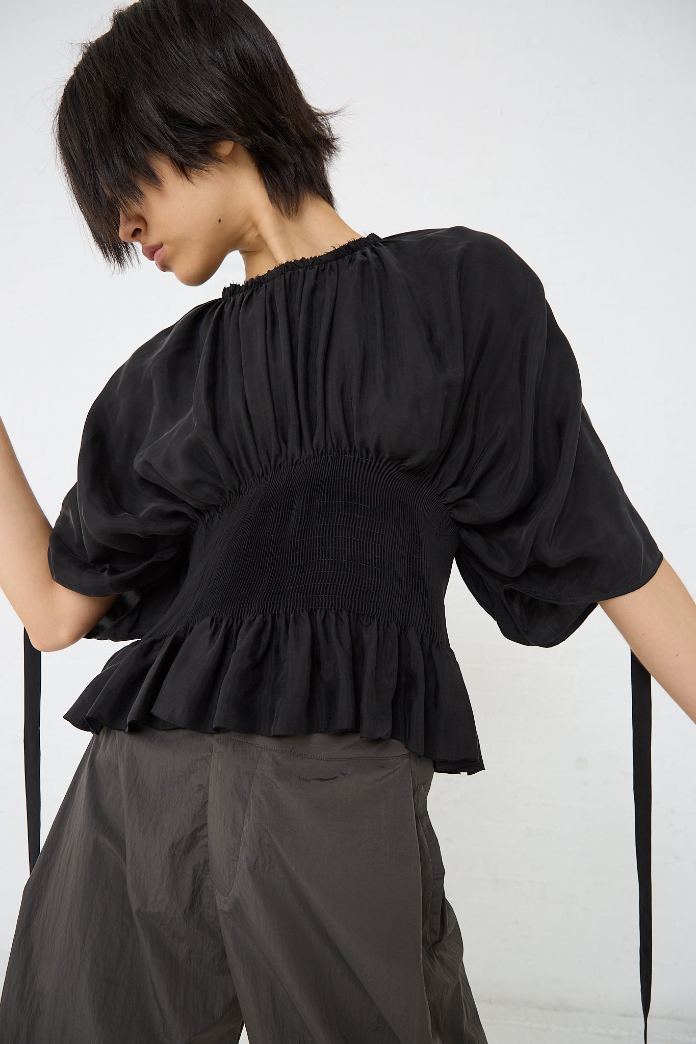 A woman wearing a Renata Brenha Cupro Sanfona Top in Washed Black with a ruffled sleeve made from recycled cupro fabric.
