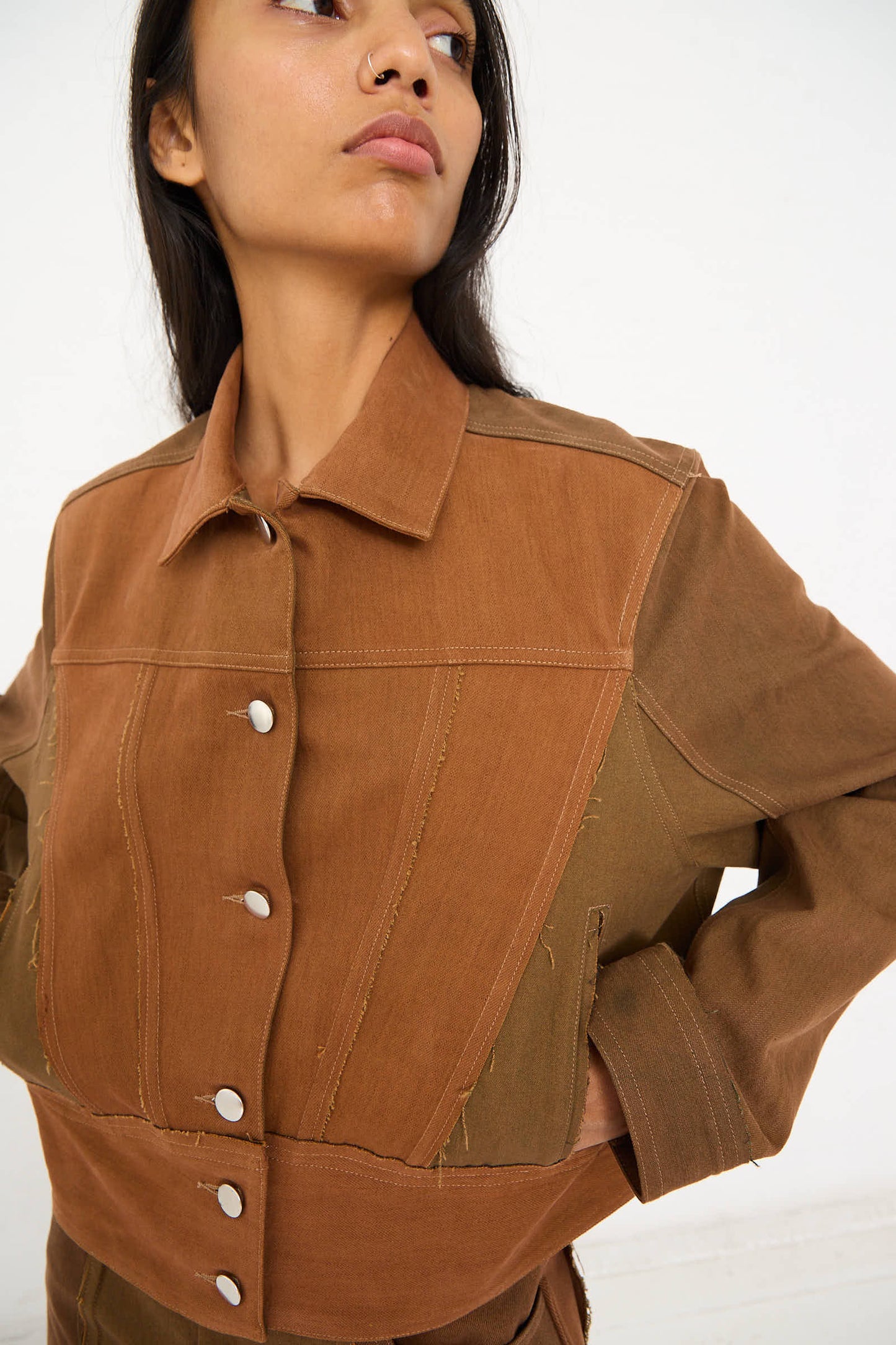 A person with long dark hair is wearing a relaxed fit, Upcycled Denim Cangaço Jacket in Brown by Renata Brenha. The two-tone brown cotton jacket features button closures and frayed edges. They are pictured from the torso up, looking off to the side.