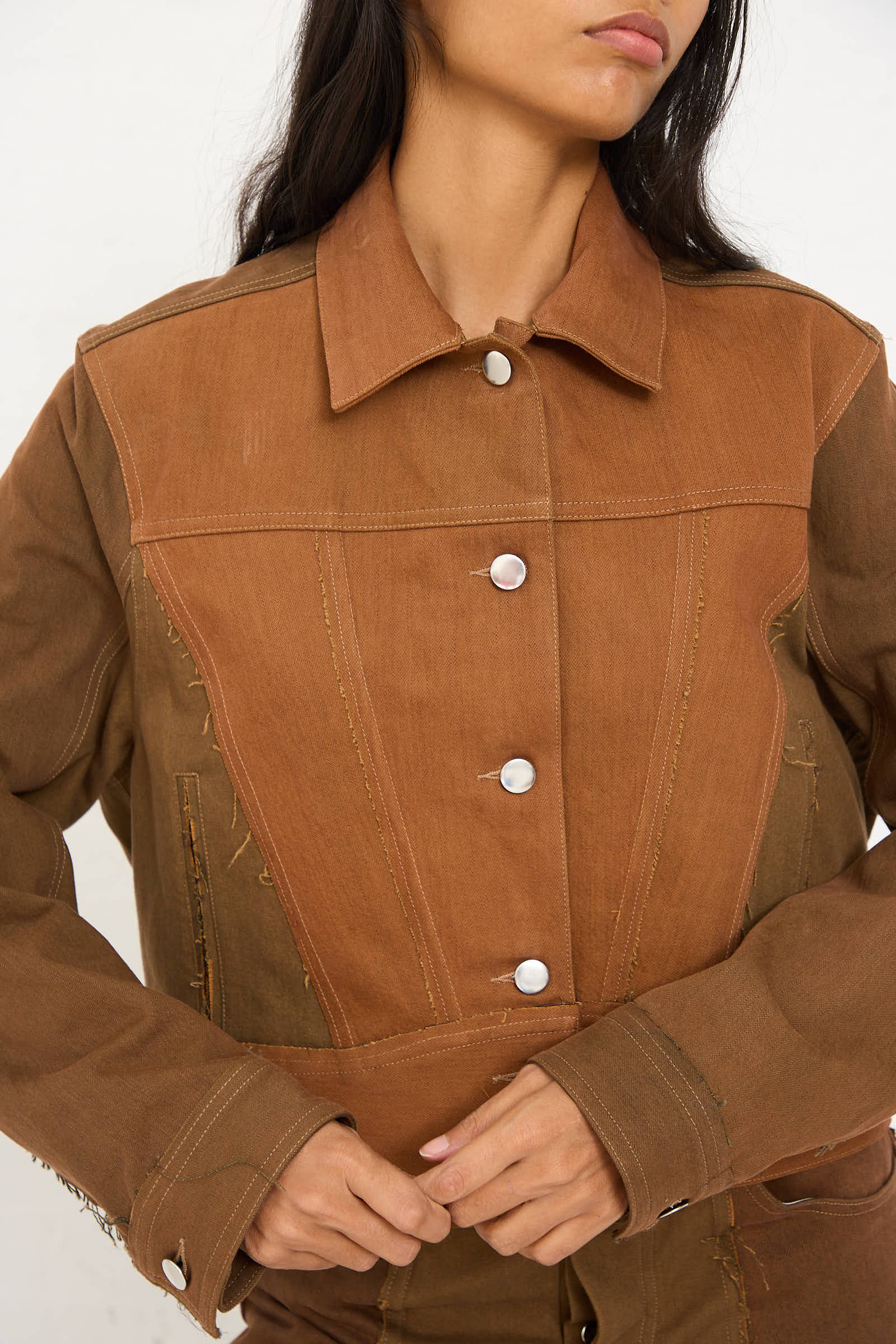 A woman wearing the Renata Brenha Upcycled Denim Cangaço Jacket in Brown, featuring a relaxed fit, with hands resting at the bottom sides. The jacket showcases slight variations in brown shades and prominent stitching details.