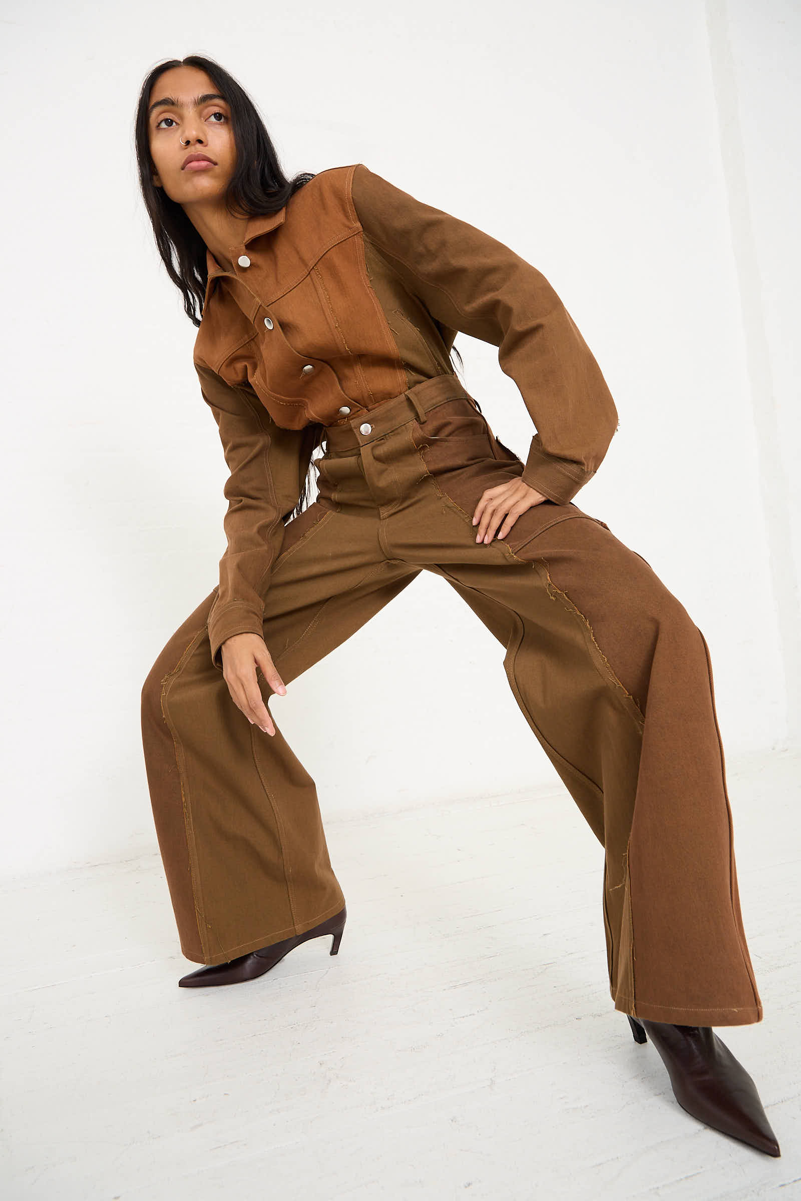 A woman with long dark hair poses in Renata Brenha's Upcycled Denim Cangaço Trouser in Brown, a stylish two-piece outfit featuring a jacket and wide-leg pants with zipper details. The relaxed-fit jacket perfectly complements her oversized trousers. She wears dark pointed-toe shoes and stands against a white background.