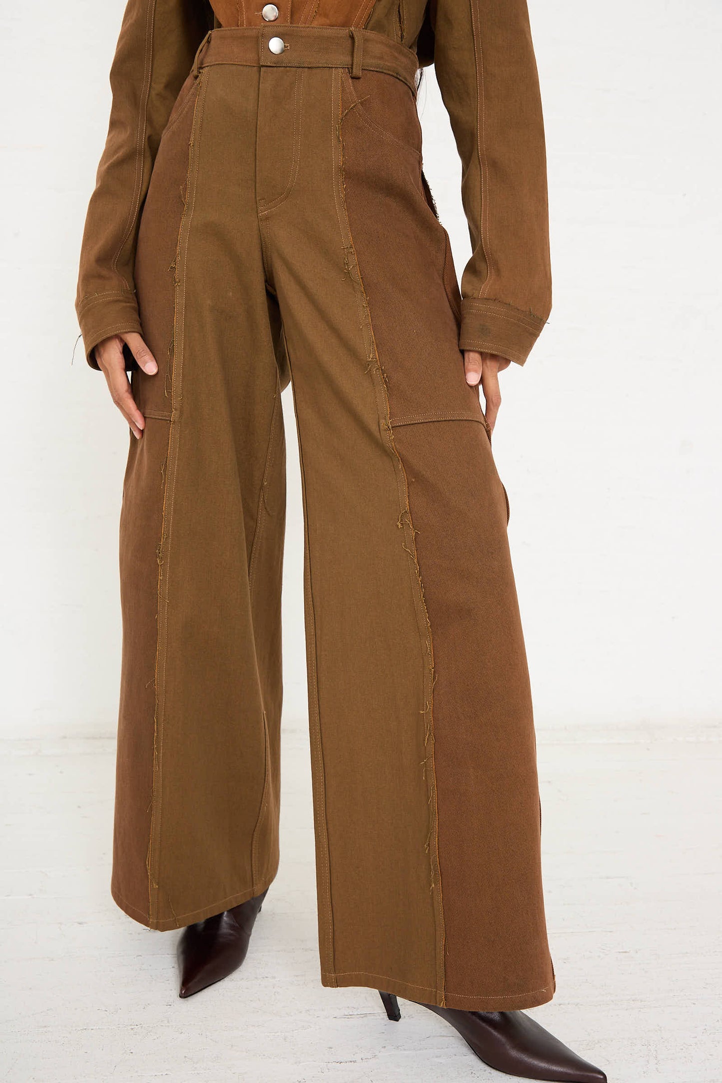 A person wearing Renata Brenha's Upcycled Denim Cangaço Trouser in Brown, a matching brown jacket, and pointed-toe boots. The high-waisted, wide-leg trousers with visible seams and a relaxed fit add to their casual elegance. Only the lower body is visible.