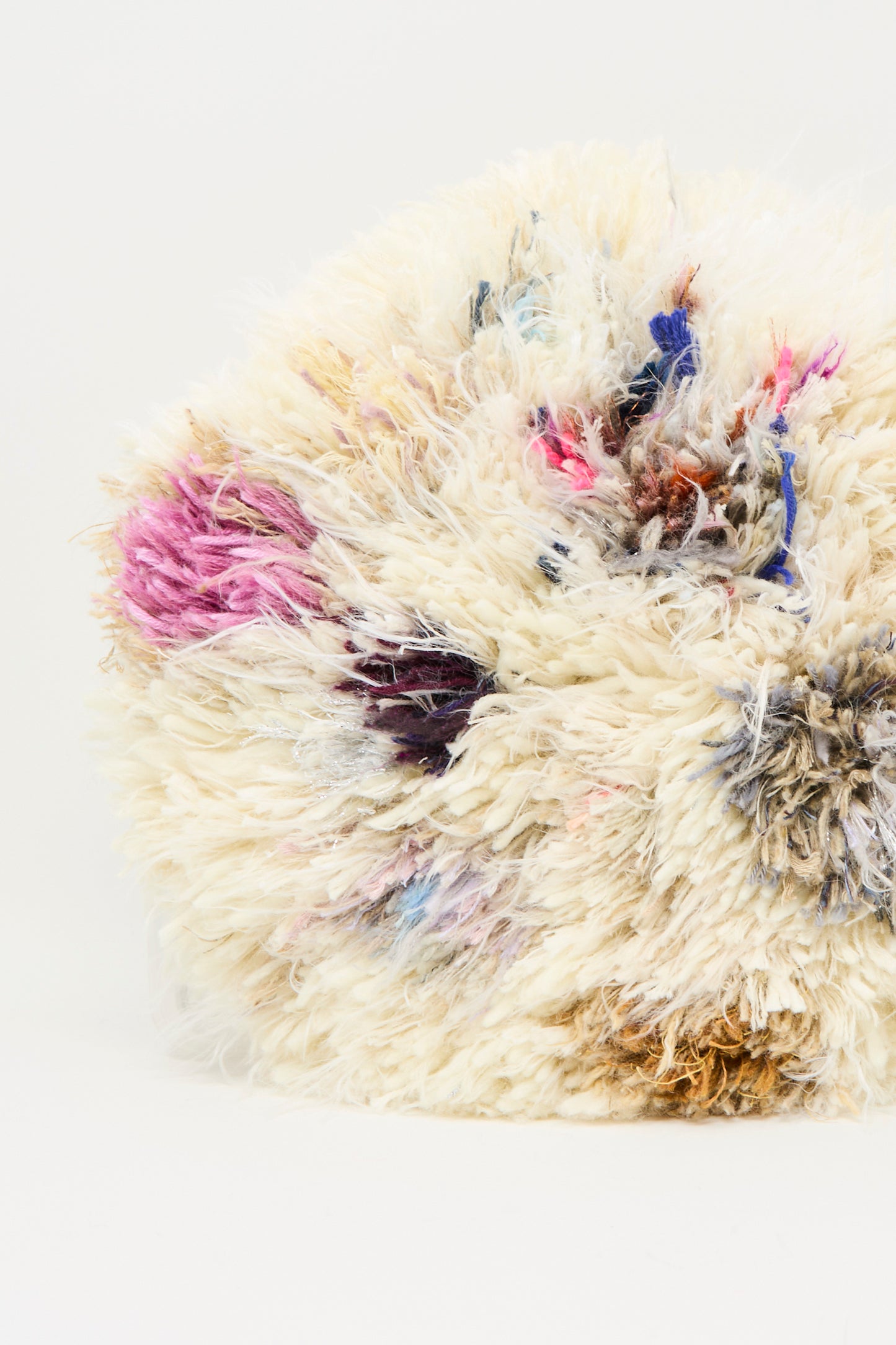 A close-up of the Handmade Cushion in Multi III by Renilde De Peuter, a round, fluffy seat cushion featuring off-white, pink, blue, black, and brown fibers against a plain white background.