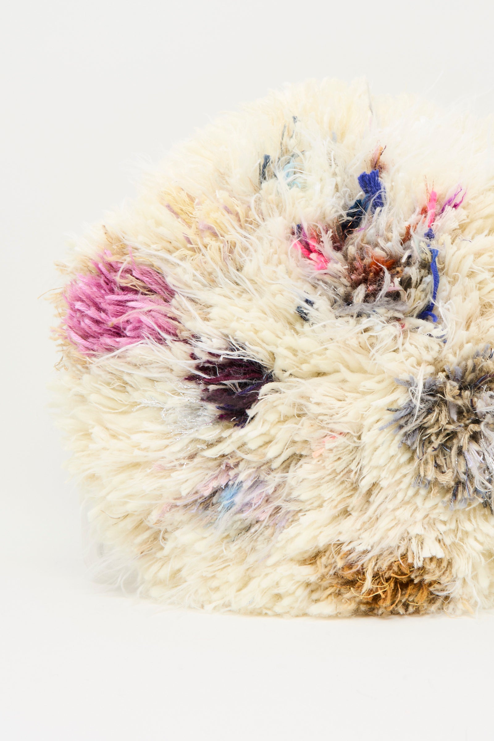 A close-up of the Handmade Cushion in Multi III by Renilde De Peuter, a round, fluffy seat cushion featuring off-white, pink, blue, black, and brown fibers against a plain white background.
