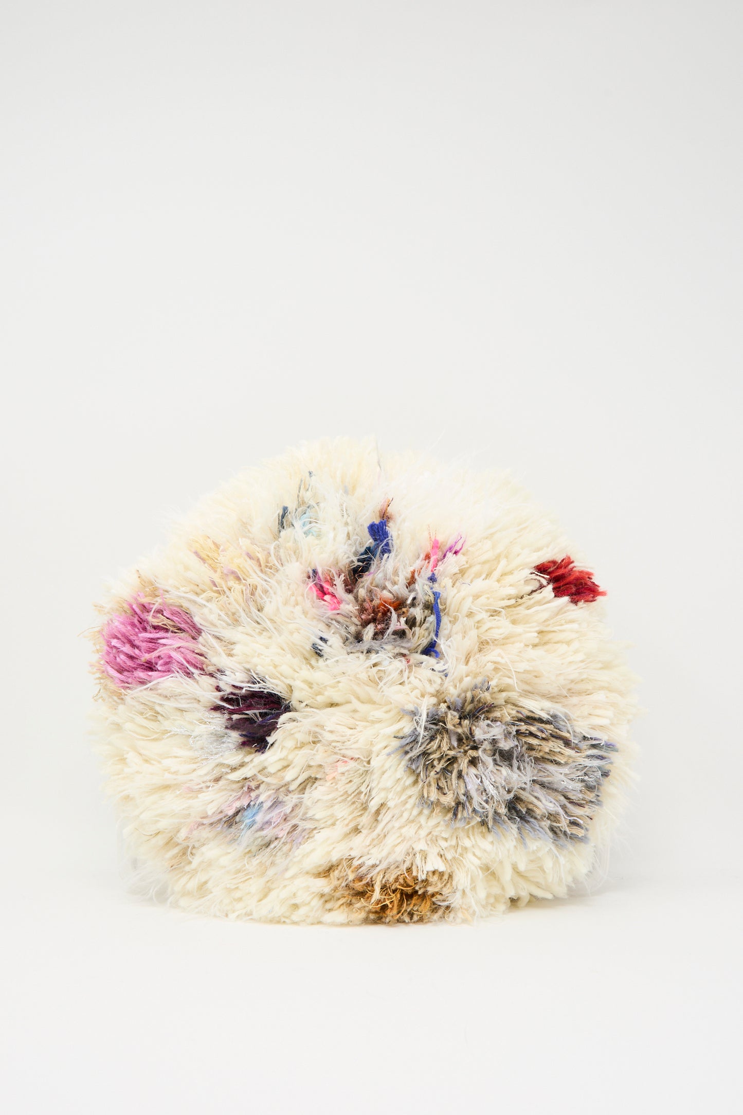 The Handmade Cushion in Multi III by Renilde De Peuter is a round, fluffy seat cushion with an off-white shaggy texture and multicolored patches of pink, blue, red, and gray on a plain white background.