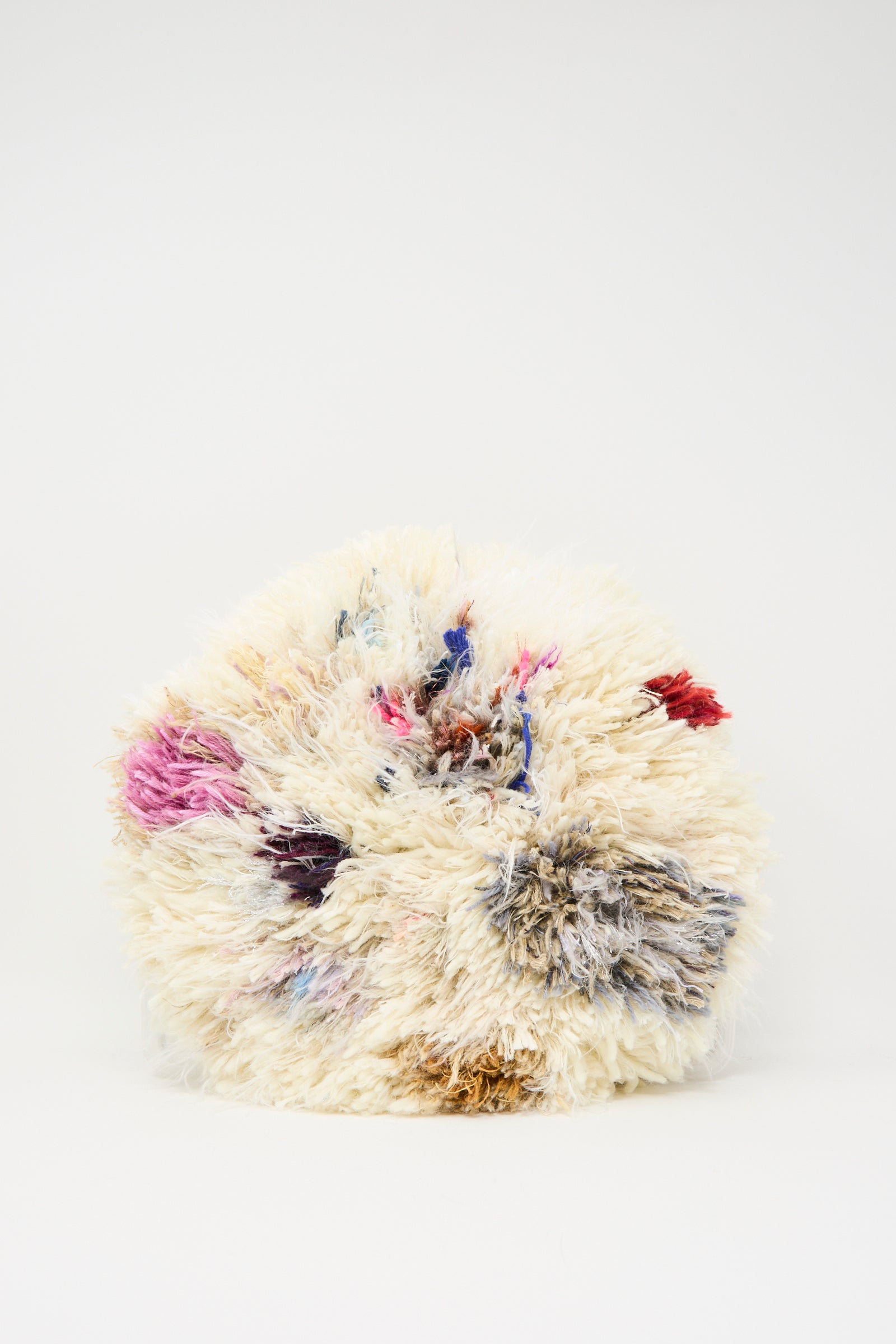 The Handmade Cushion in Multi III by Renilde De Peuter is a round, fluffy seat cushion with an off-white shaggy texture and multicolored patches of pink, blue, red, and gray on a plain white background.