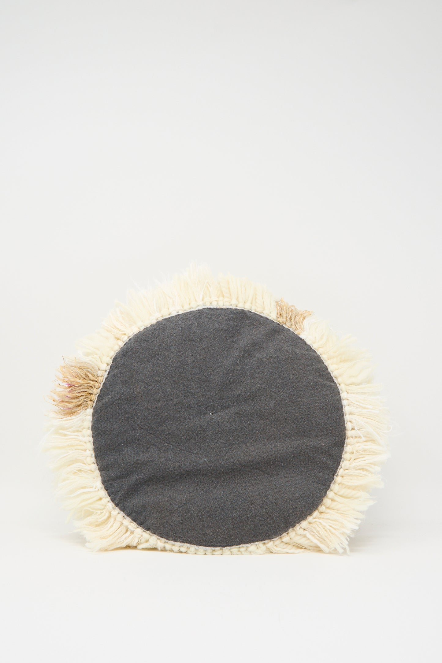 The Handmade Cushion in Multi III by Renilde De Peuter features a round shape with a black fabric center and cream fringe edges, smyrna stitch-inspired, shown on a plain white background.