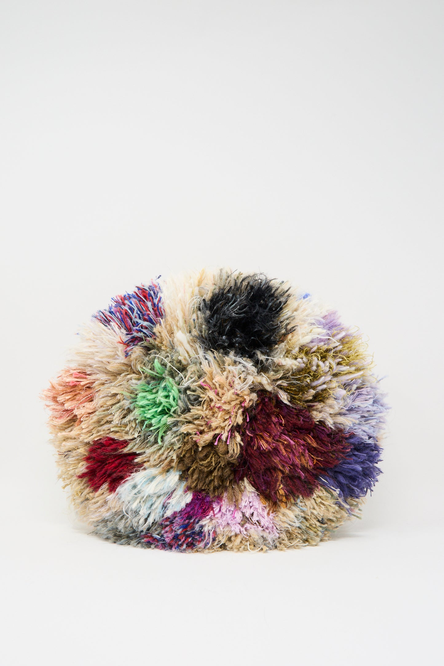 The Handmade Cushion in Multi V by Renilde De Peuter is a round, fluffy cushion crafted from recycled, multi-colored yarns and displayed against a plain white background.