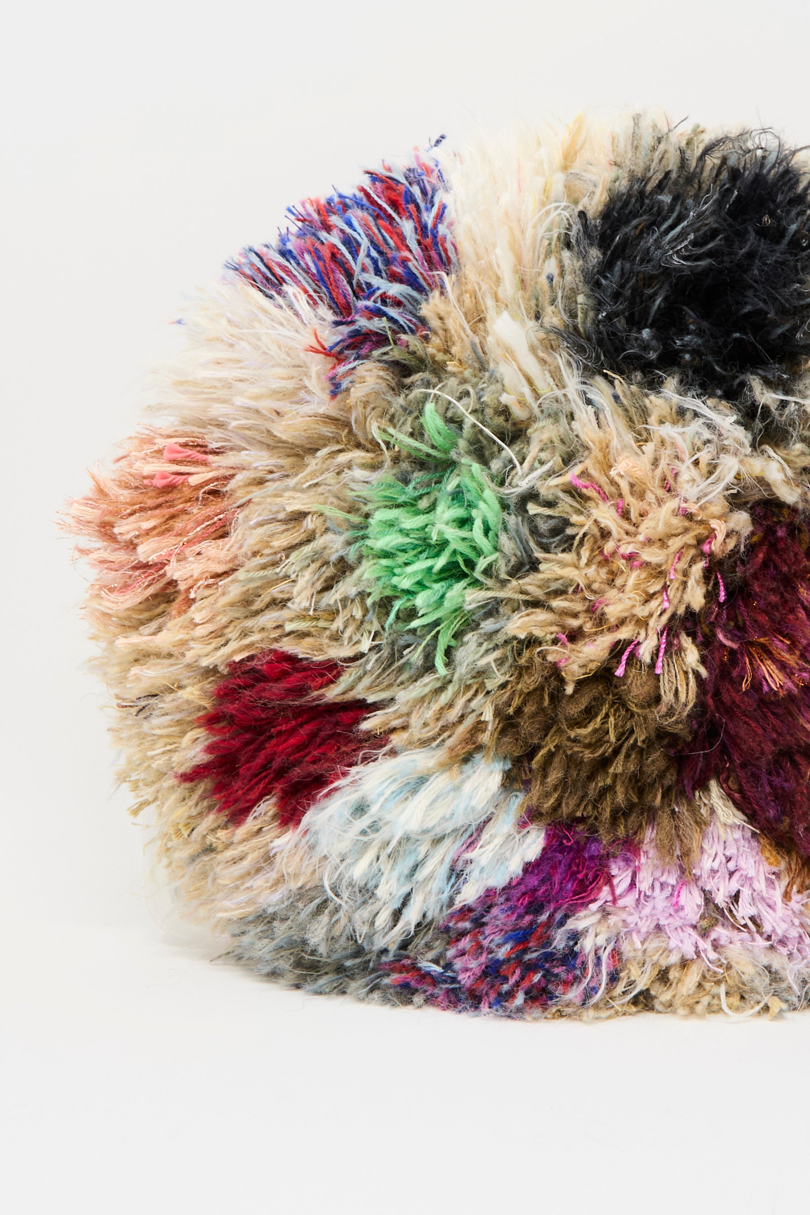 A close-up of the Handmade Cushion in Multi V by Renilde De Peuter, a shaggy, round seat cushion made from recycled yarns with vibrant patches, showcased against a plain white background.