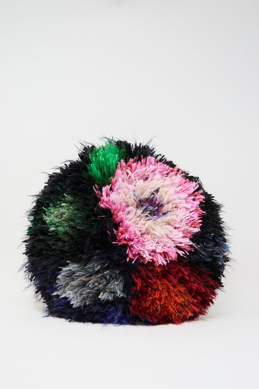 The Handmade Cushion in Multi XIV by Renilde De Peuter features a round, shaggy design made from colorful patches of green, pink, black, gray, red, and blue fabrics. It is shown against a plain white background.