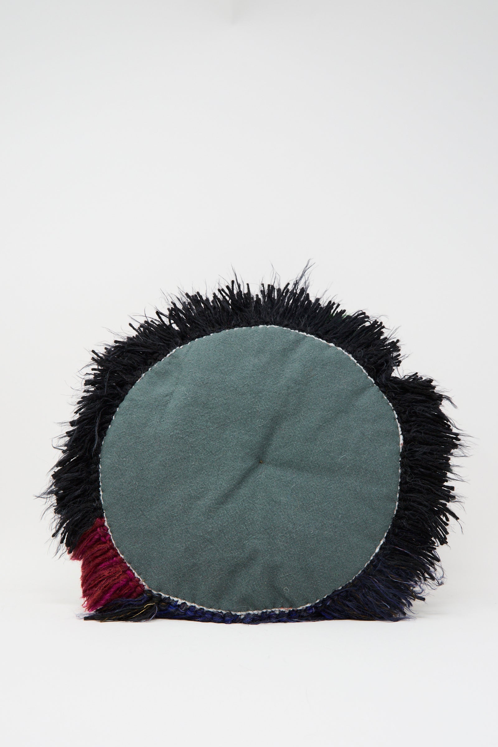 The Handmade Cushion in Multi XIV by Renilde De Peuter features a central green fabric with bold black, red, and blue fringe, crafted using the smyrna stitch and displayed on a plain white background.